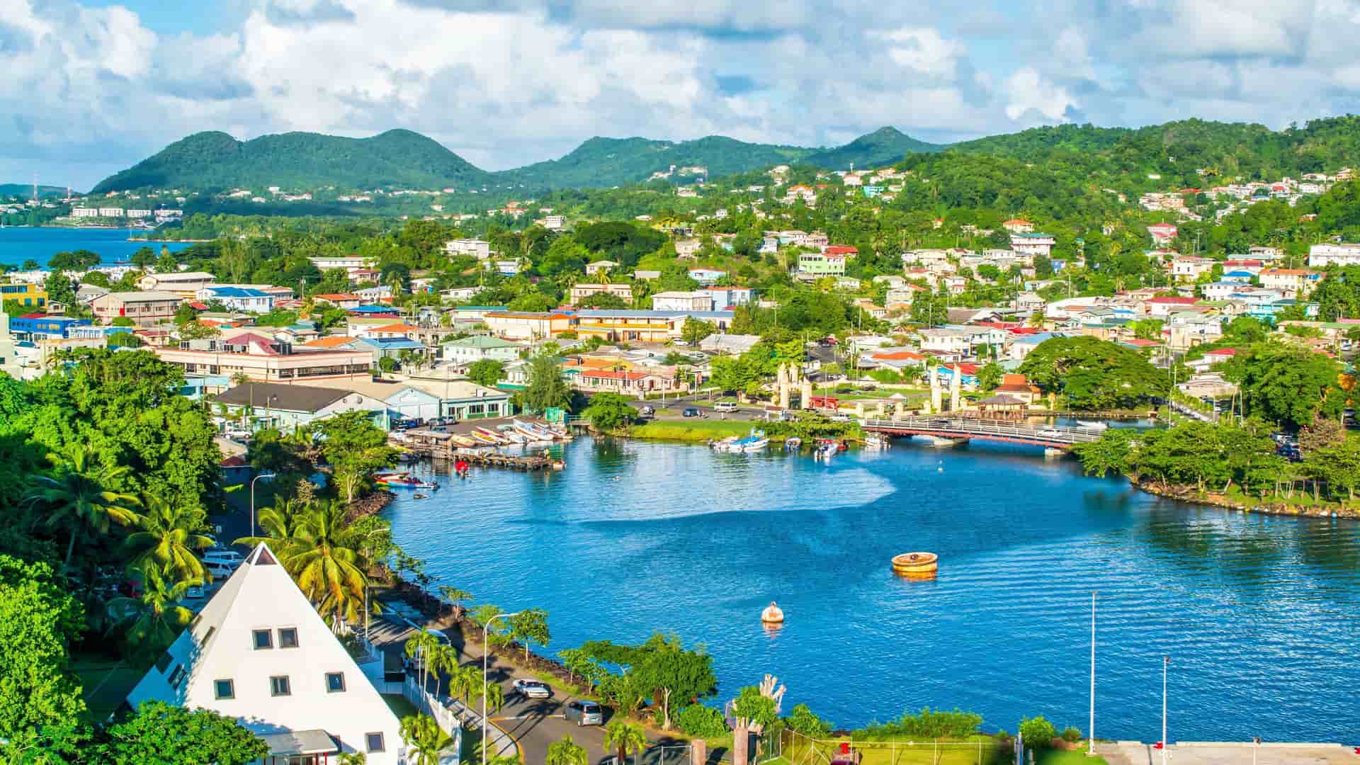 A vibrant panoramic view of Castries, St. Lucia, with colorful buildings nestled along the harbor and lush green hills in the background under a blue sky with white clouds.