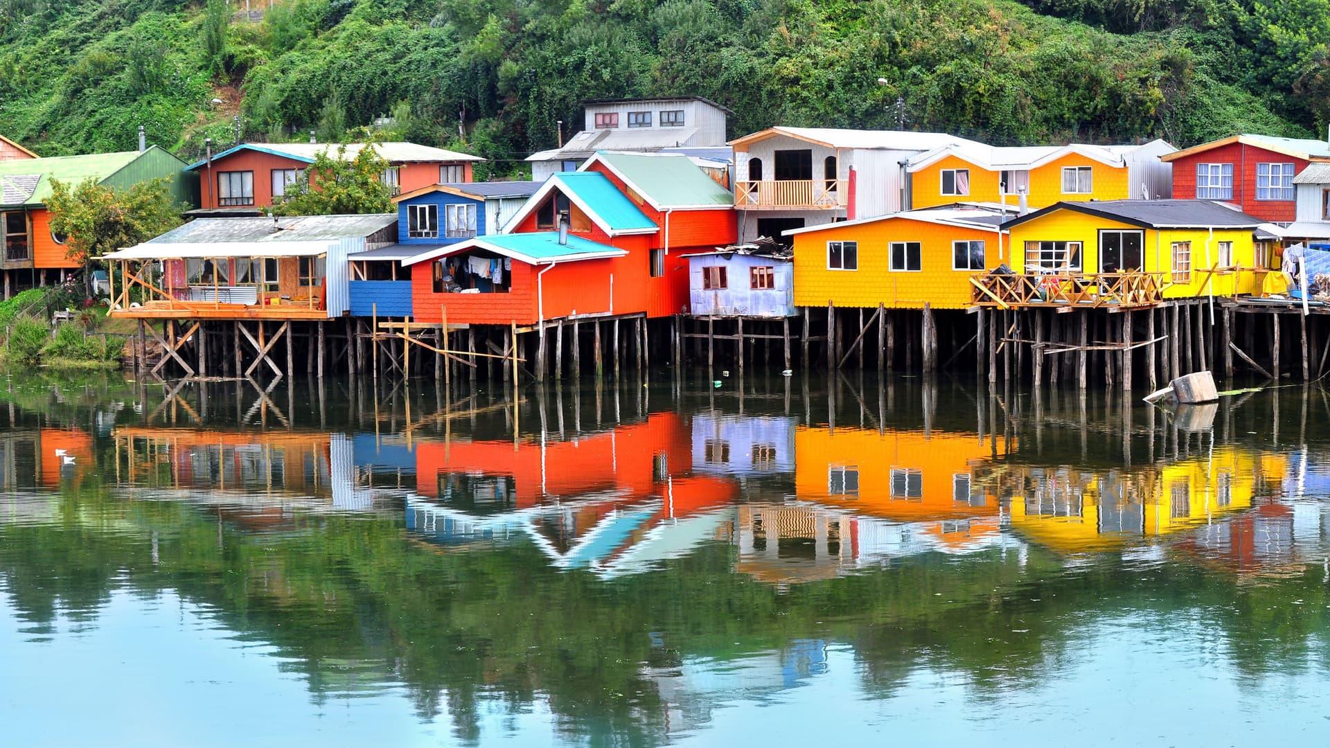 A close-up view of the colorful stilt houses, known as palafitos, lining the waterfront of Castro, Chiloé Island, Chile, with their reflections visible in the calm water.