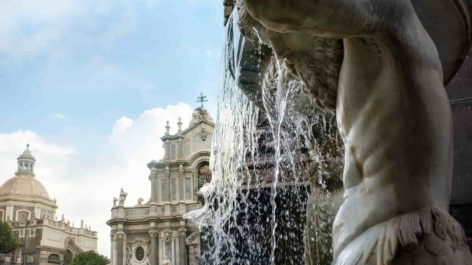 A low-angle view of the Fountain of the Elephant in Catania, Sicily, with water cascading over the sculpted body of a mythical figure and the dome of the Catania Cathedral in the background.