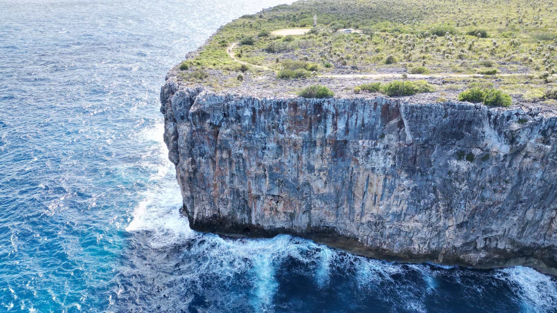 A close-up aerial view of the dramatic limestone bluff, known as the "Bluff," a landmark on Cayman Brac island, with waves crashing against its vertical face.