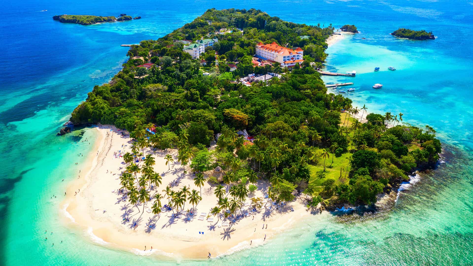 A tropical island scene in Cayo Levantado with white sandy beaches, turquoise water, palm trees, and colorful resort buildings, with visitors relaxing on the shore and boats anchored near the coastline.