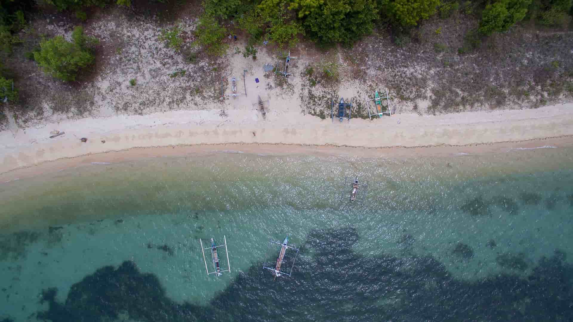 An aerial shot of Celukan Bawang beach in Bali, Indonesia, with clear, shallow water and traditional fishing boats, known as jukungs, moored near the shoreline.