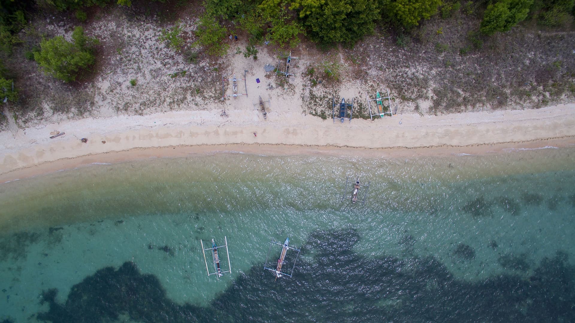 An aerial shot of Celukan Bawang beach in Bali, Indonesia, with clear, shallow water and traditional fishing boats, known as jukungs, moored near the shoreline.