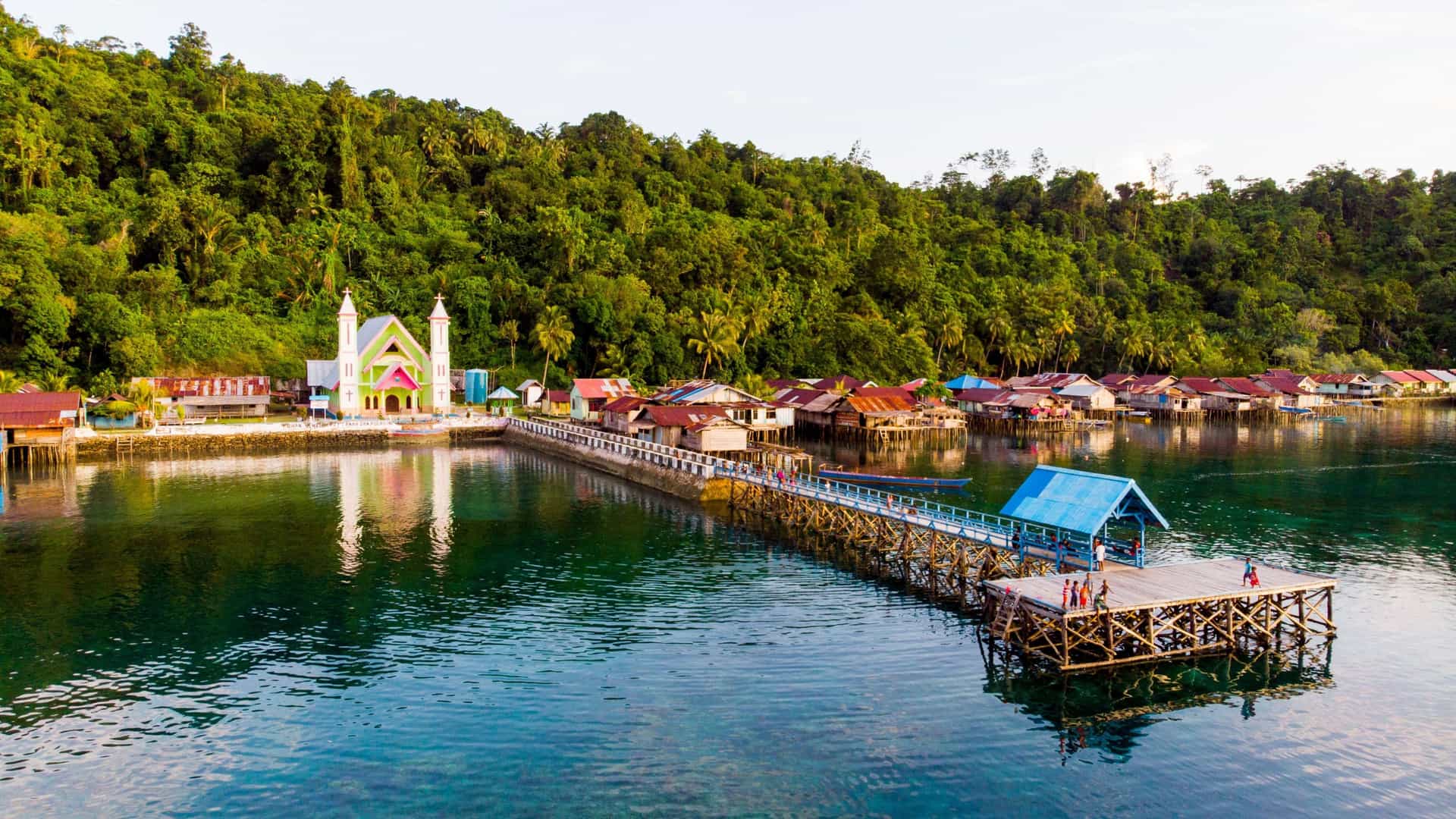 An aerial view of a vibrant stilt village on the coast of Cenderawasih Bay, with a colorful church and houses built over the water, backed by lush green jungle.
