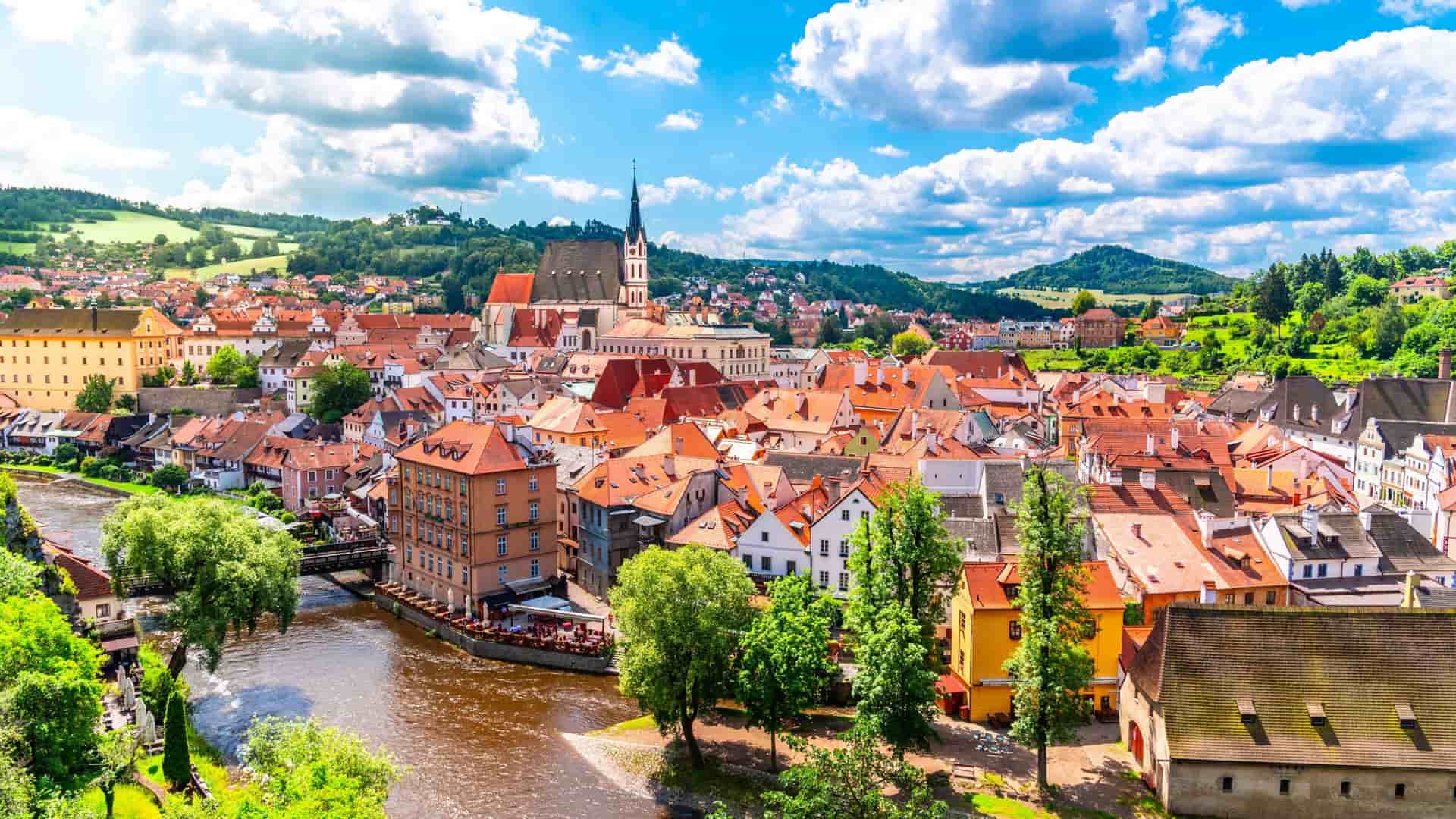 A vibrant panoramic view of the medieval town of Cesky Krumlov, with its red-roofed buildings, a winding river, and a prominent church steeple against a blue, cloudy sky.