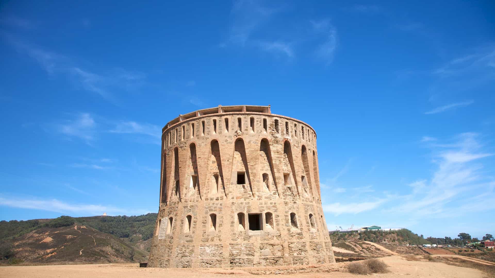 A low-angle shot of the historic Molino del Dique tower in Ceuta, a round, stone structure with arched openings, set against a bright blue sky with hills in the background.