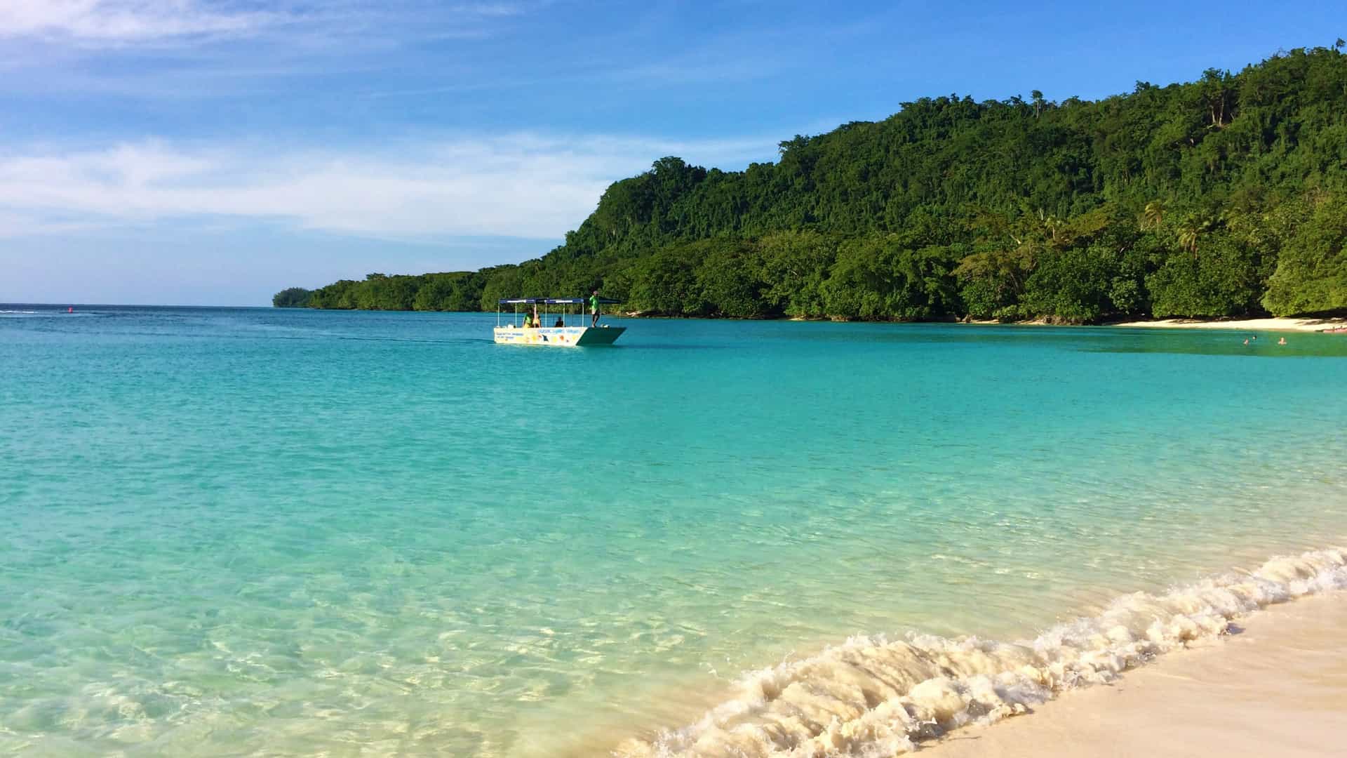 Clear turquoise waves lap against the white sand of Champagne Bay, with a small boat anchored near a dense, jungle-covered hillside.