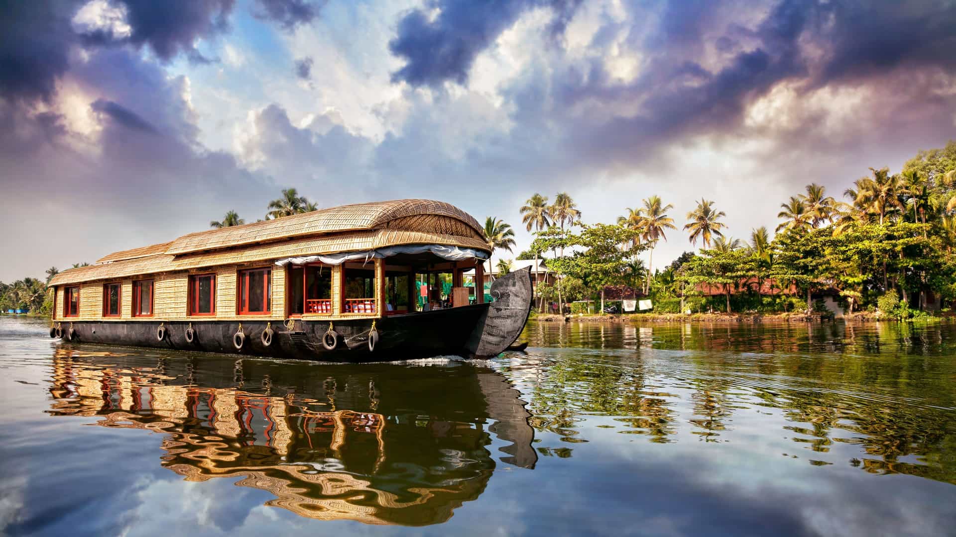 A beautiful floating houseboat with a thatched roof cruising along a canal in the backwaters of Chandannagar, India, surrounded by lush tropical trees and a cloudy sky.