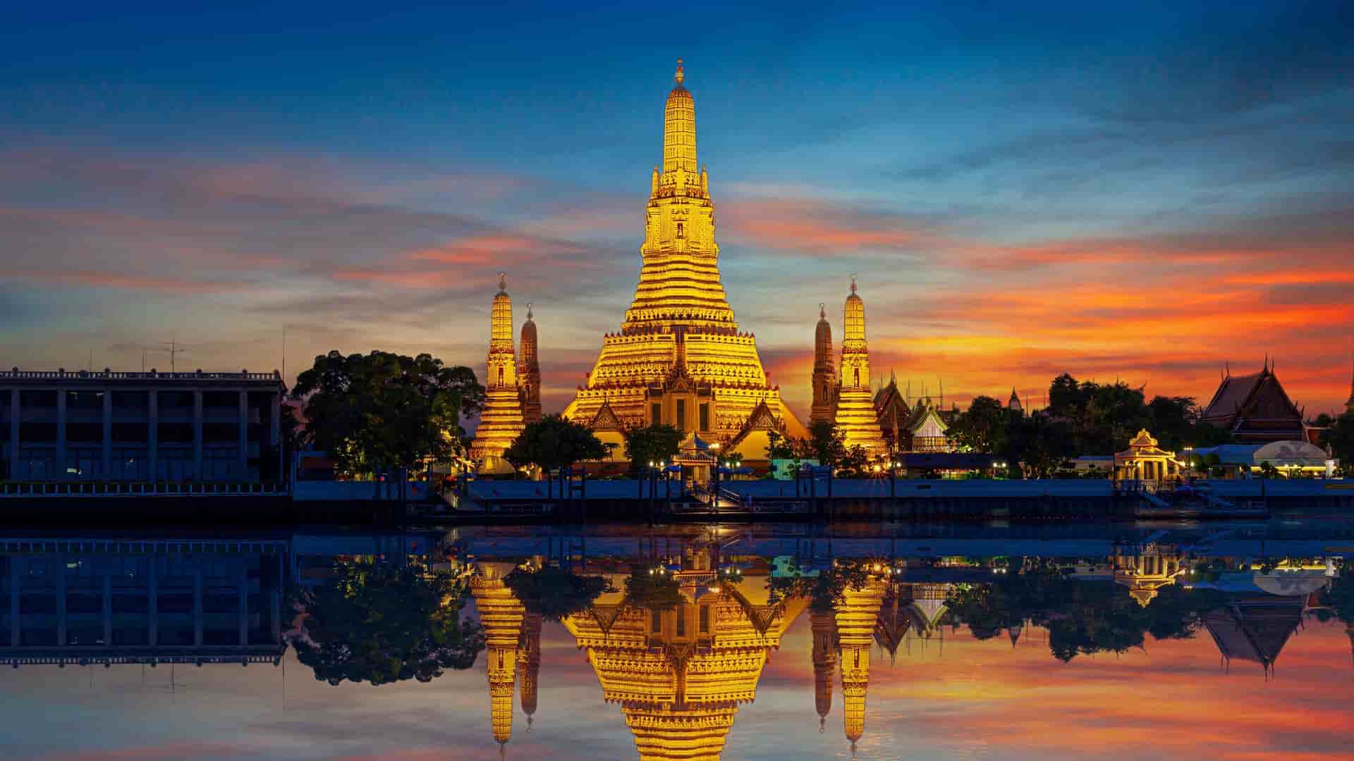 A vibrant sunset view of the illuminated Wat Arun temple, known as the Temple of Dawn, reflecting on the surface of the Chao Phraya River in Bangkok, Thailand.
