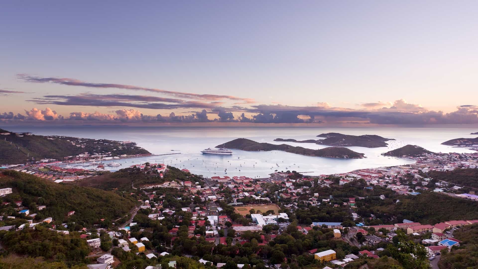 A breathtaking panoramic view of Charlotte Amalie harbor in St. Thomas, with a cruise ship anchored in the bay, surrounded by rolling green hills and a colorful sunset.