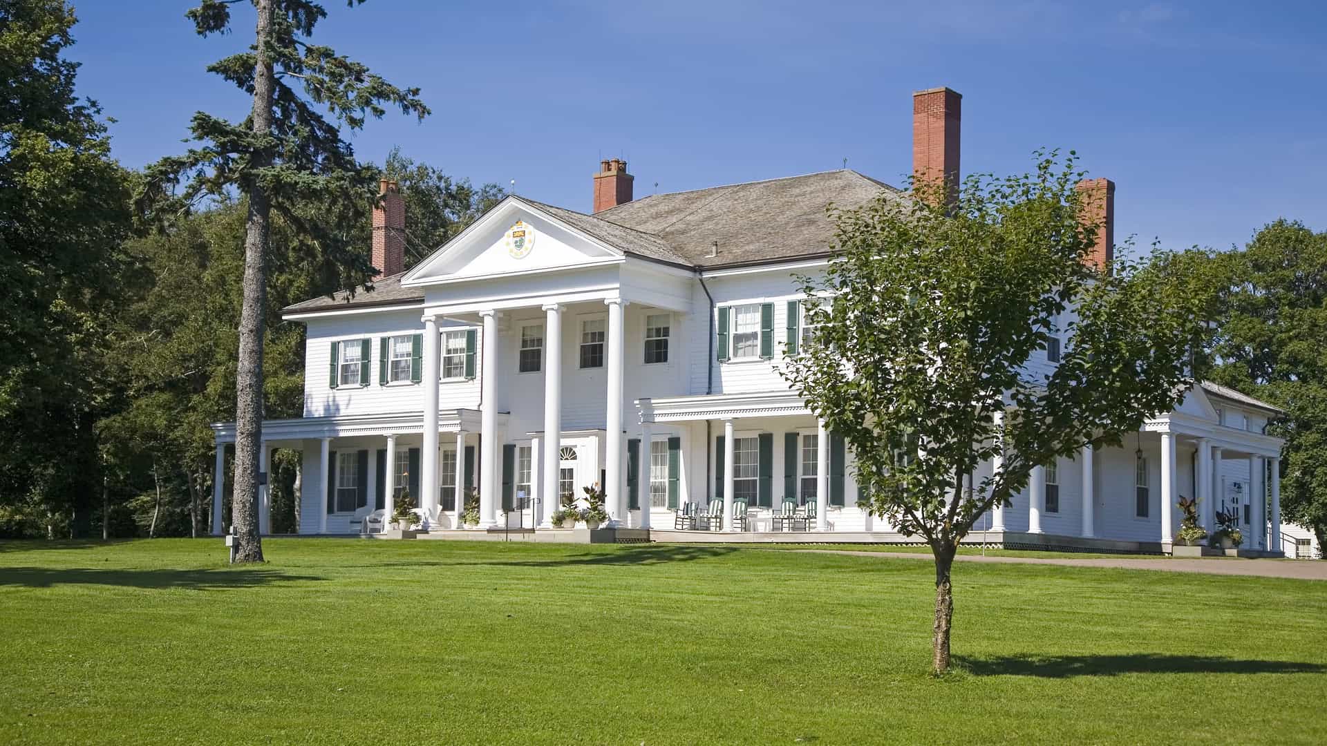 A wide shot of the Government House, a large white mansion with a Greek Revival facade and a prominent portico, surrounded by a green lawn and tall trees in Charlottetown.