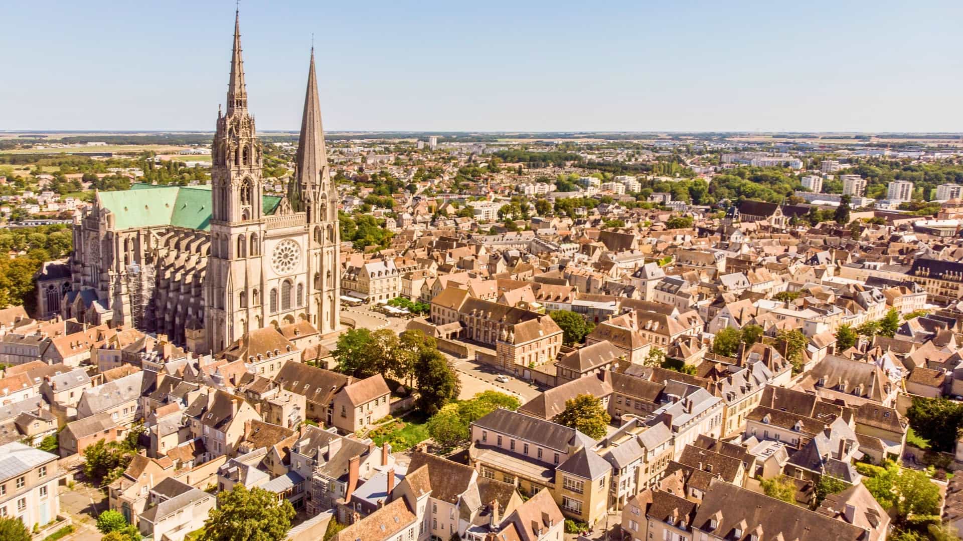 An aerial view of the historic city of Chartres, France, with the spires and flying buttresses of the magnificent Notre-Dame de Chartres Cathedral rising above the rooftops.