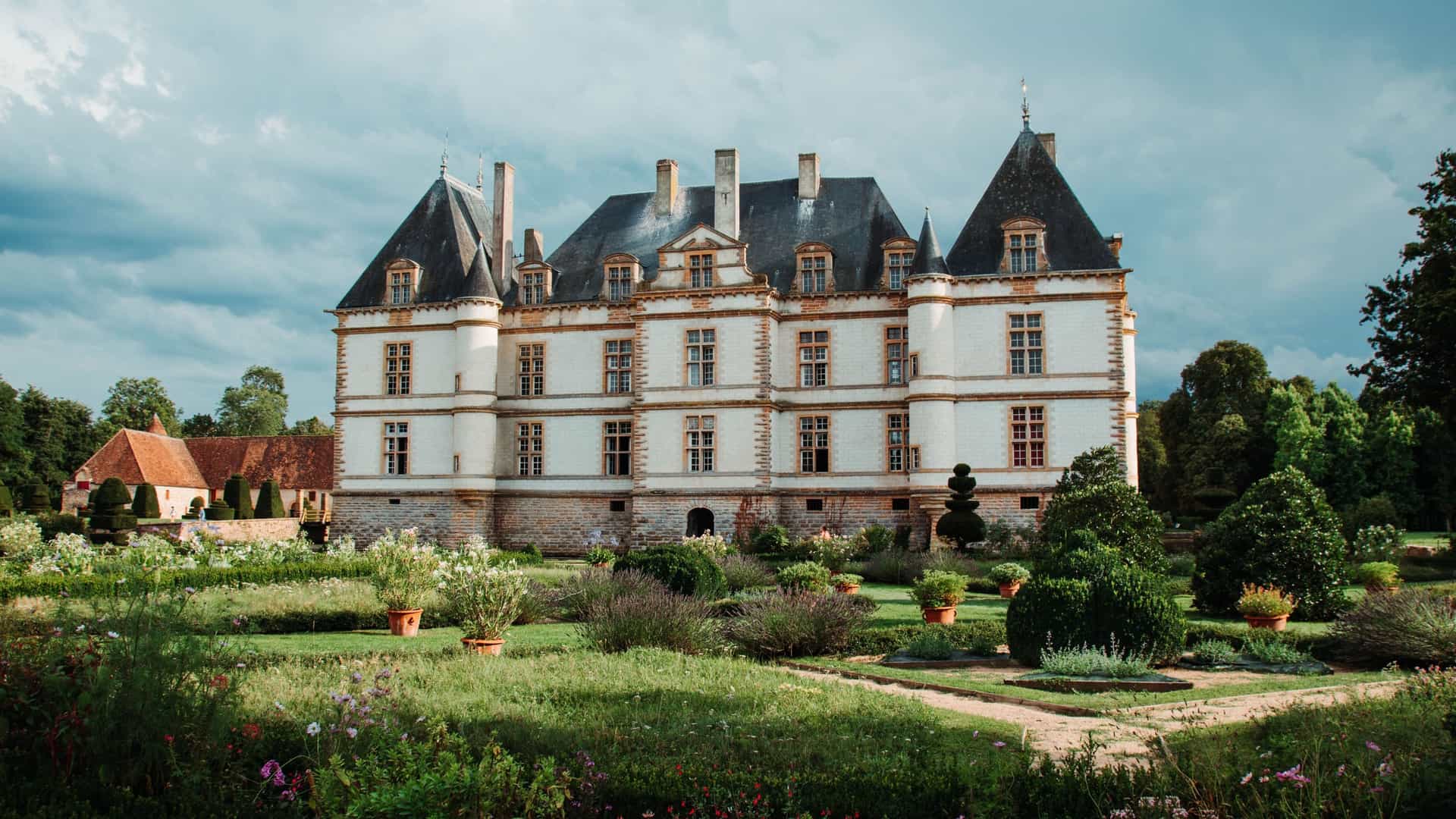 A full view of the elegant Château de Cormatin, a beautiful 17th-century castle with white walls and slate roofs, surrounded by manicured gardens under a cloudy sky.