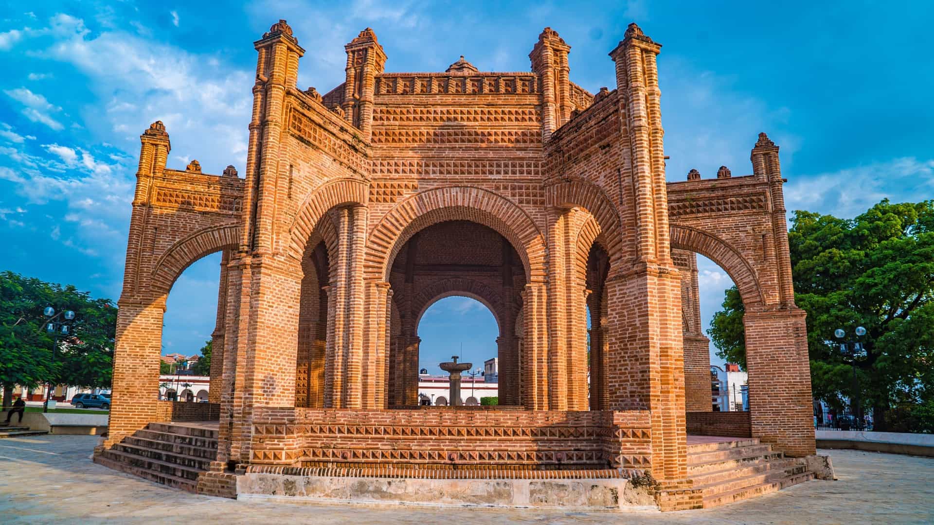 A historic brick structure with multiple arches and a fountain visible through the center, in a sunny plaza in Chiapas, Mexico.