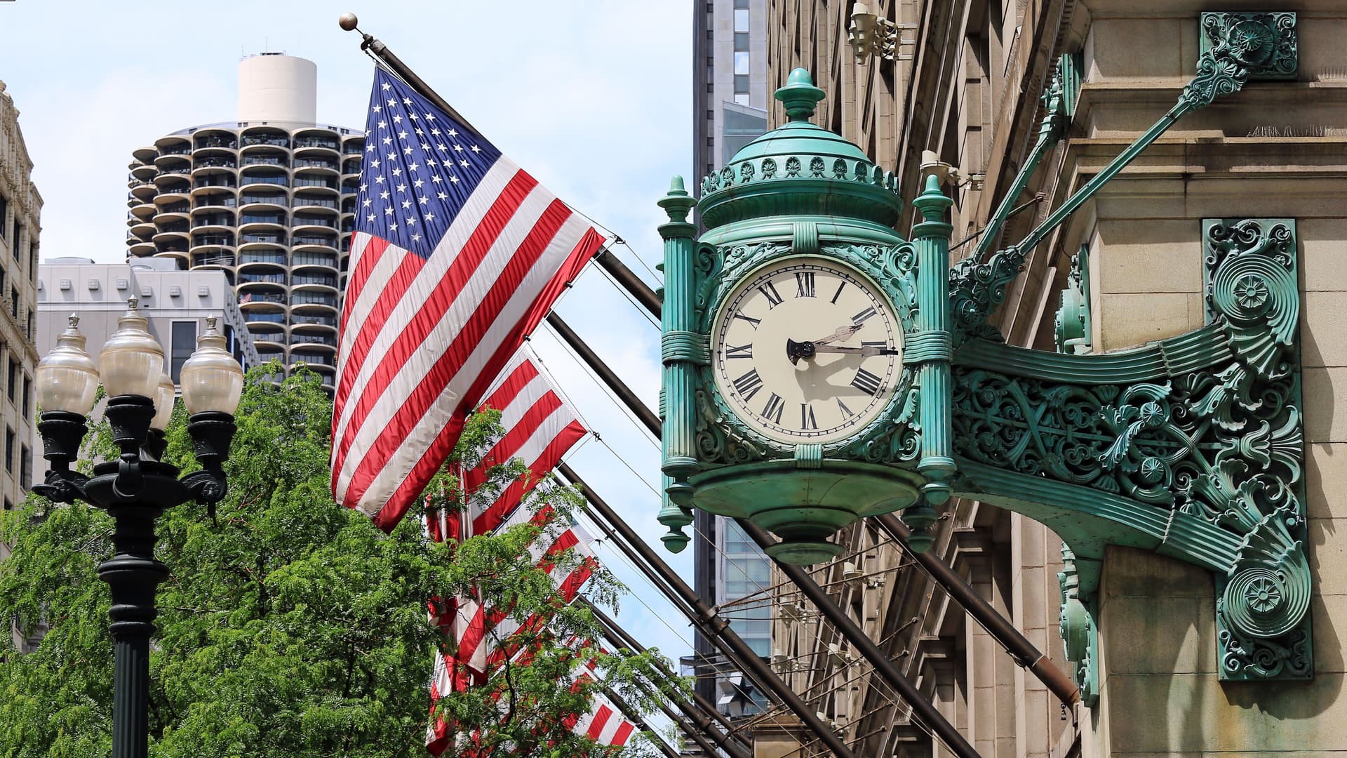A historic green-patina clock on a building in Chicago, with multiple American flags and city lights in the background.