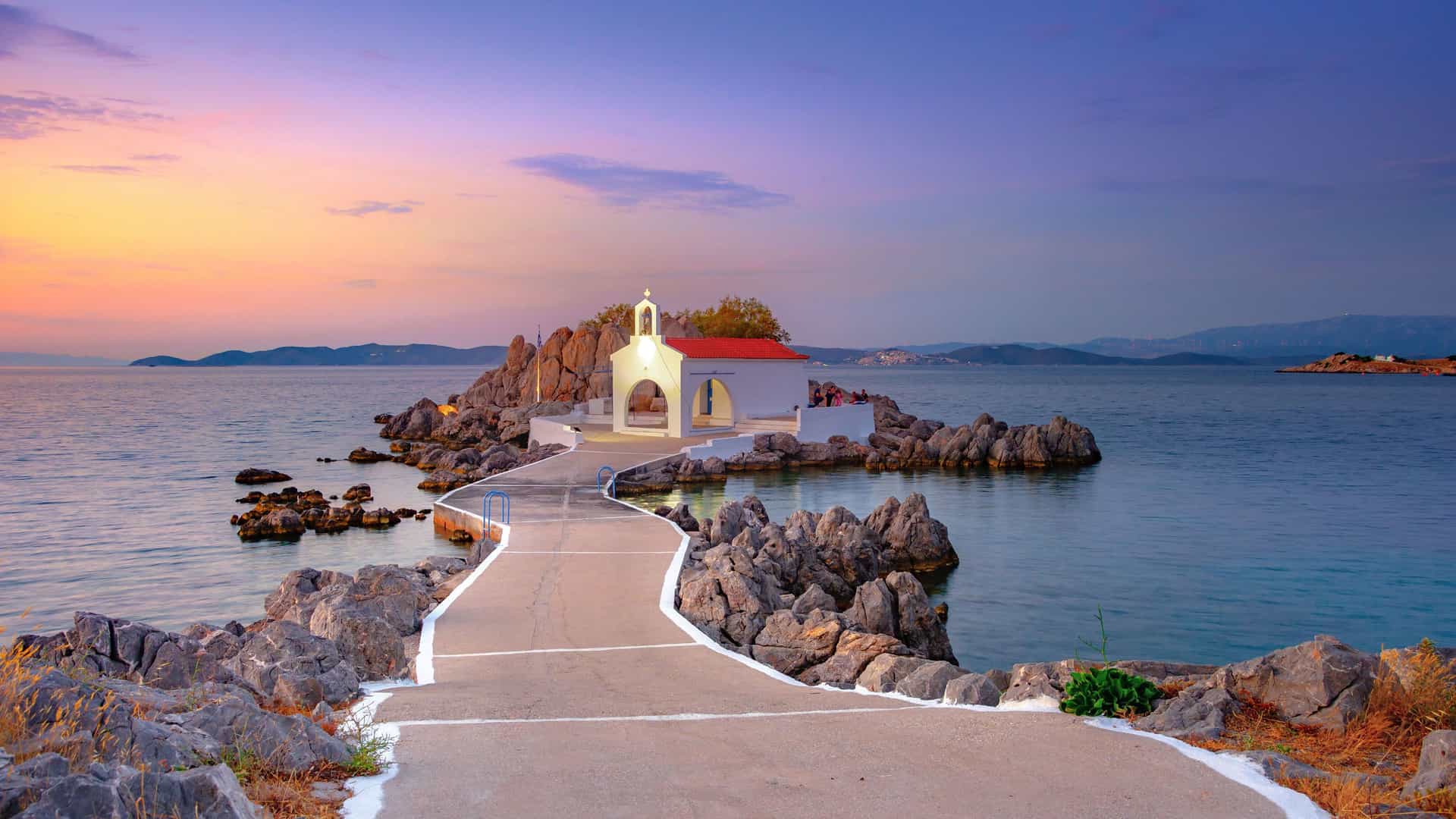 A picturesque seaside church on a rocky islet connected by a paved walkway to the mainland, with a vibrant sunset sky over the Aegean Sea in Chios, Greece.