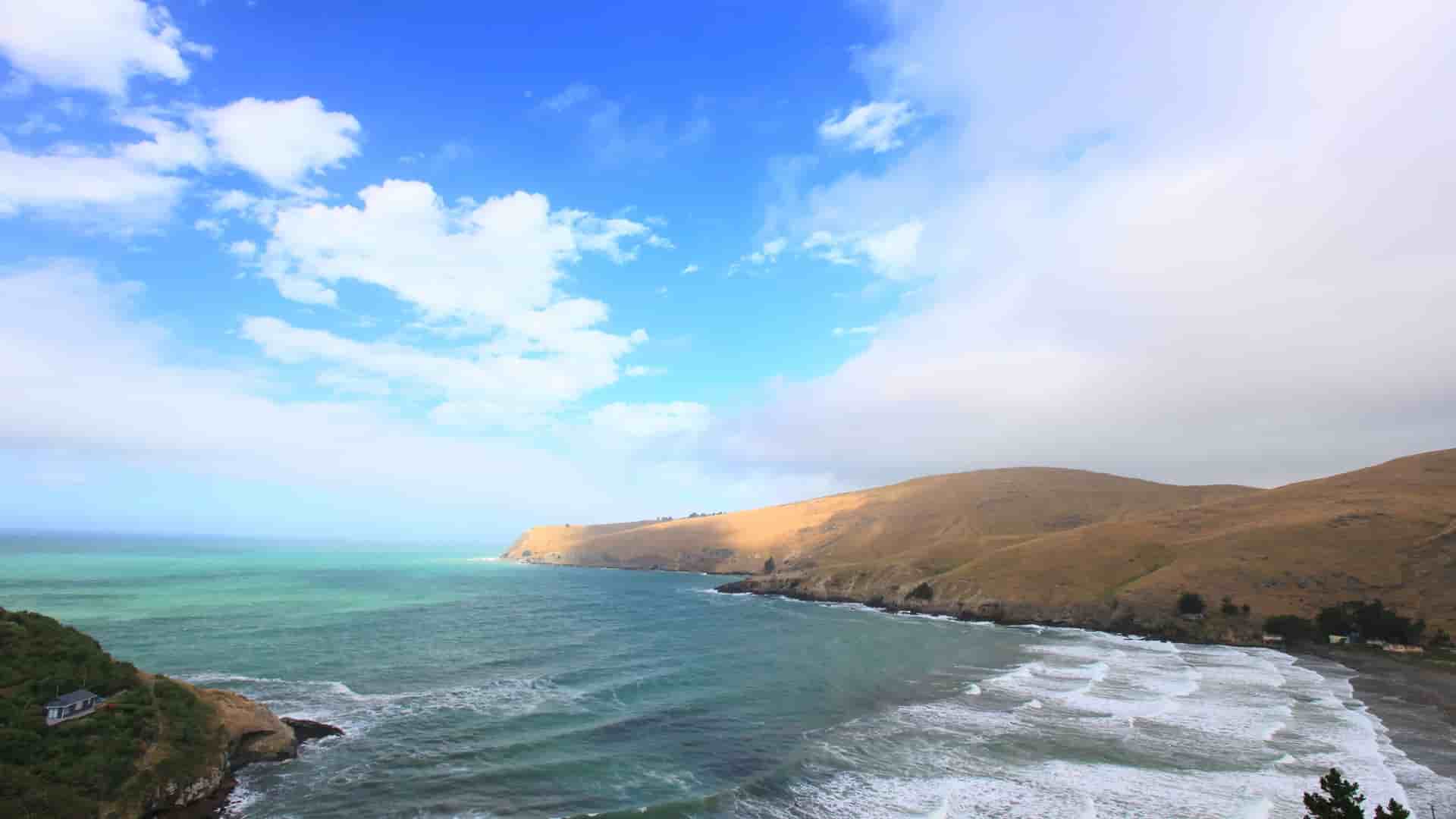 A panoramic view of the dramatic, rolling hills and cliffs overlooking a turquoise bay with waves, on the coastline near Christchurch, New Zealand.