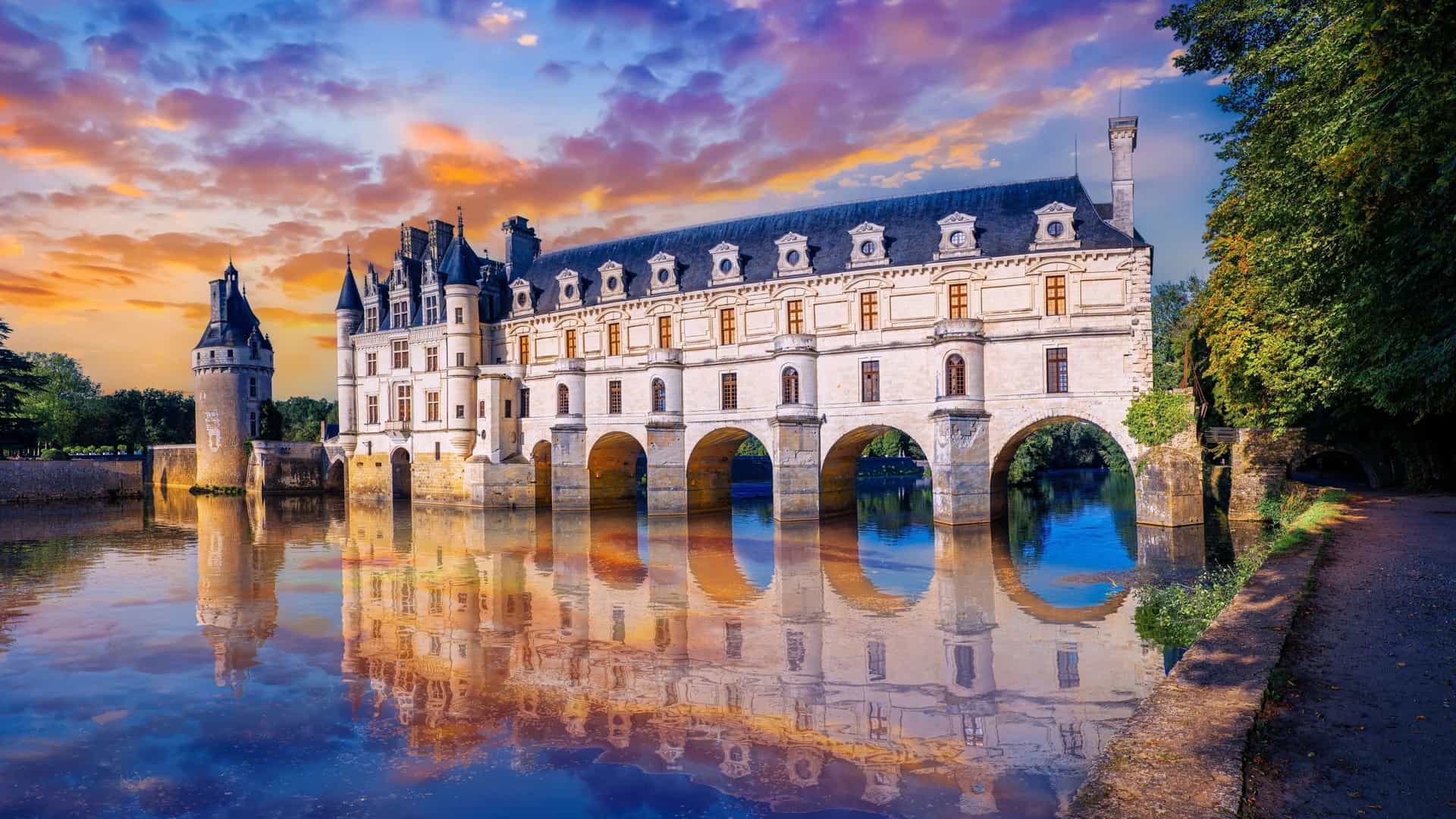 A breathtaking sunset shot of the Chateau de Chenonceau, with its distinctive gallery built over the River Cher, reflecting in the calm water with vibrant clouds.
