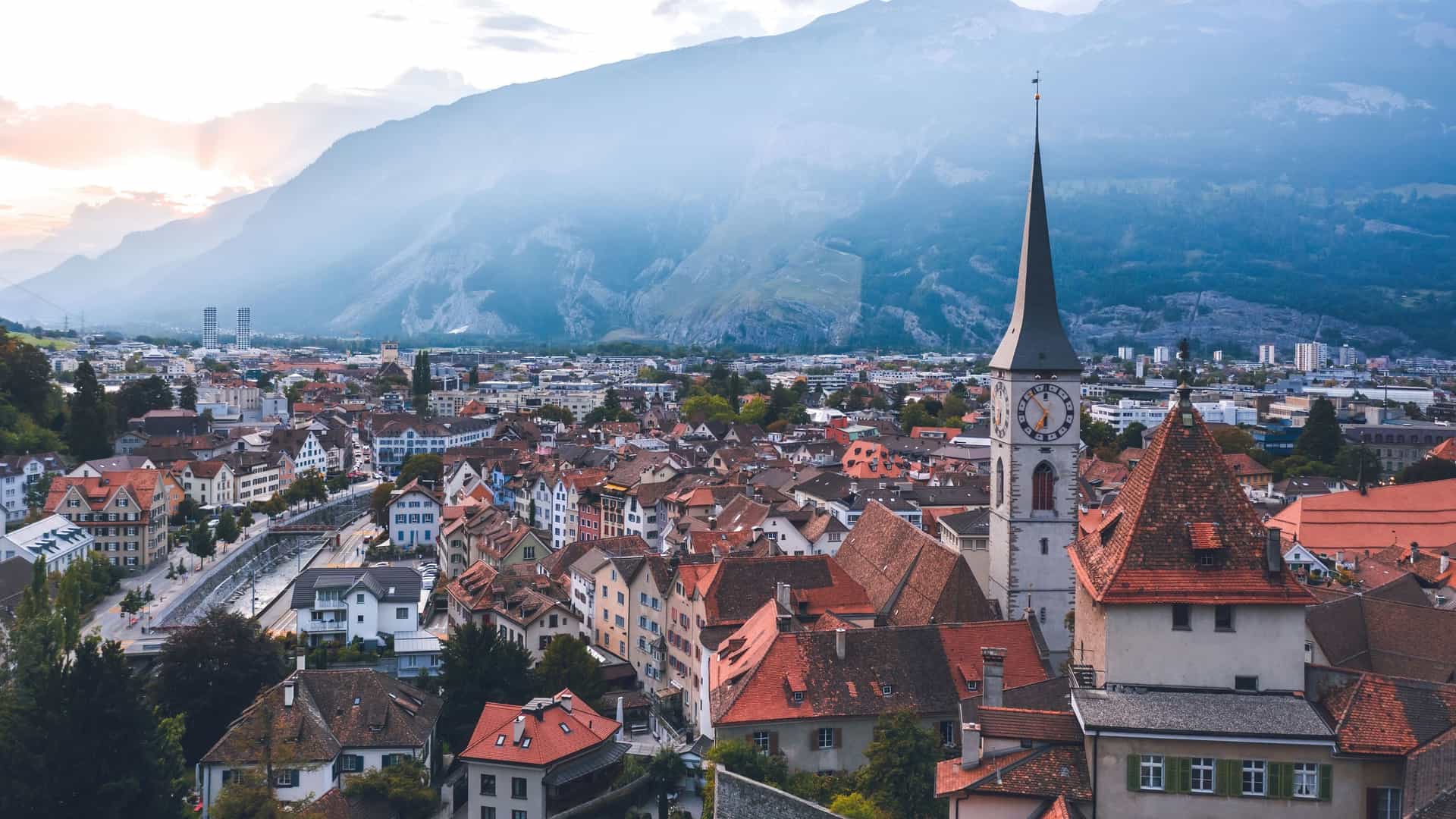 An aerial view of Chur, Switzerland, with its historic old town, including a prominent clock tower, nestled against a backdrop of hazy mountains at sunset.
