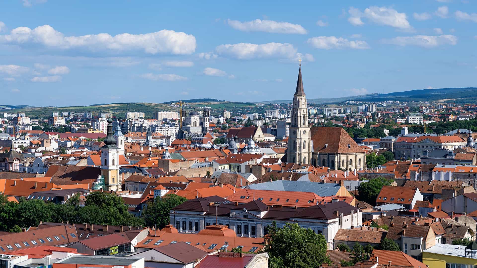 A scenic panoramic view of the old town of Cluj-Napoca, Romania, with a dense cluster of red-roofed buildings and the towering steeples of historic churches dominating the skyline, set against a backdrop of rolling green hills.