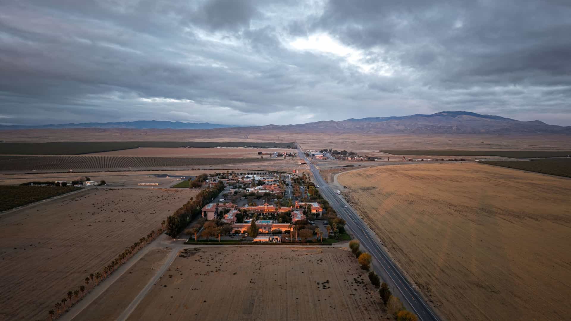 An aerial view of a hotel and small town surrounded by vast fields and a highway, with mountains in the distance, under a dramatic cloudy sky in Coalinga, California.