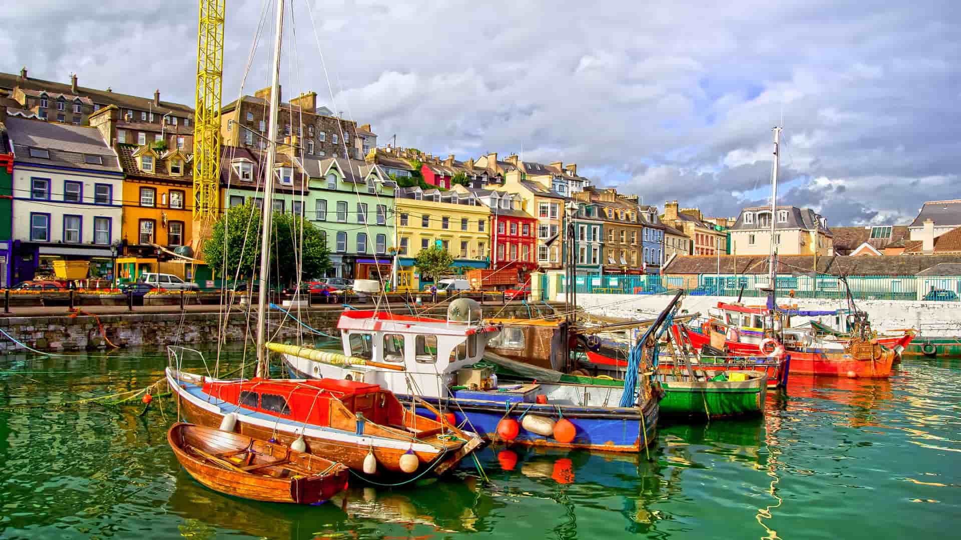 A scenic view of the colorful waterfront of Cobh, Ireland, with brightly painted houses and a variety of fishing boats docked in the harbor.