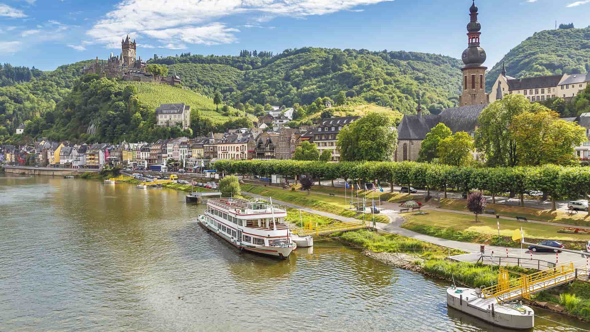 A beautiful view of Cochem, Germany, showing the historic Reichsburg Castle on a hill, the town's colorful buildings, and a cruise boat on the Moselle River.