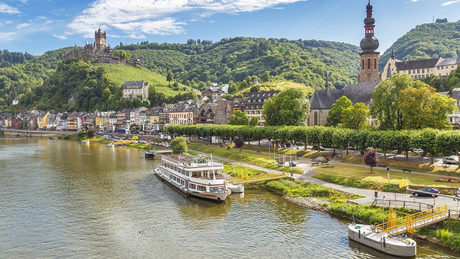 A beautiful view of Cochem, Germany, showing the historic Reichsburg Castle on a hill, the town's colorful buildings, and a cruise boat on the Moselle River.