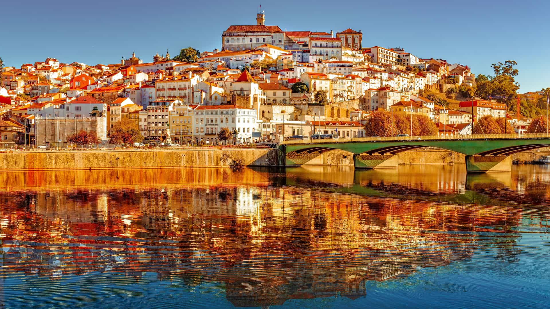 A panoramic view of the colorful city of Coimbra, Portugal, with its historic buildings and university perched on a hill, overlooking the Mondego River and a bridge.