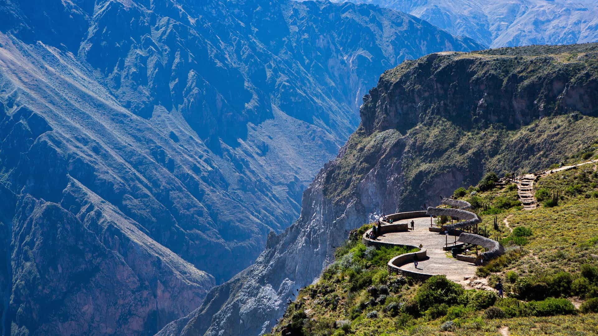 A stunning aerial view of the deep Colca Canyon in Peru, with tourists standing on a winding observation deck overlooking the dramatic, steep mountain slopes.