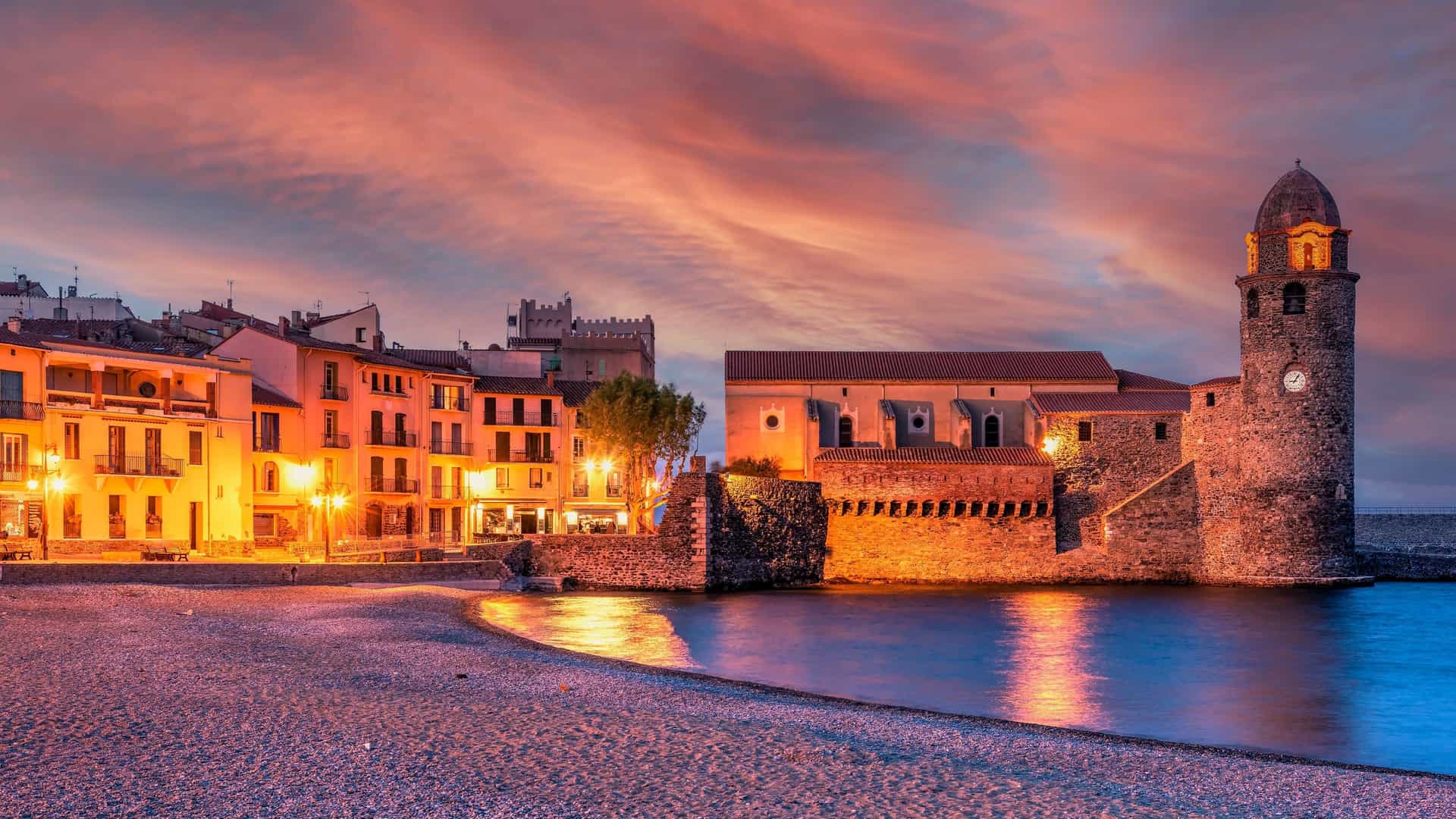 A scenic sunset over the coastal town of Collioure, France, with a prominent church and lighthouse on the right and colorful buildings lining a sandy beach at dusk.