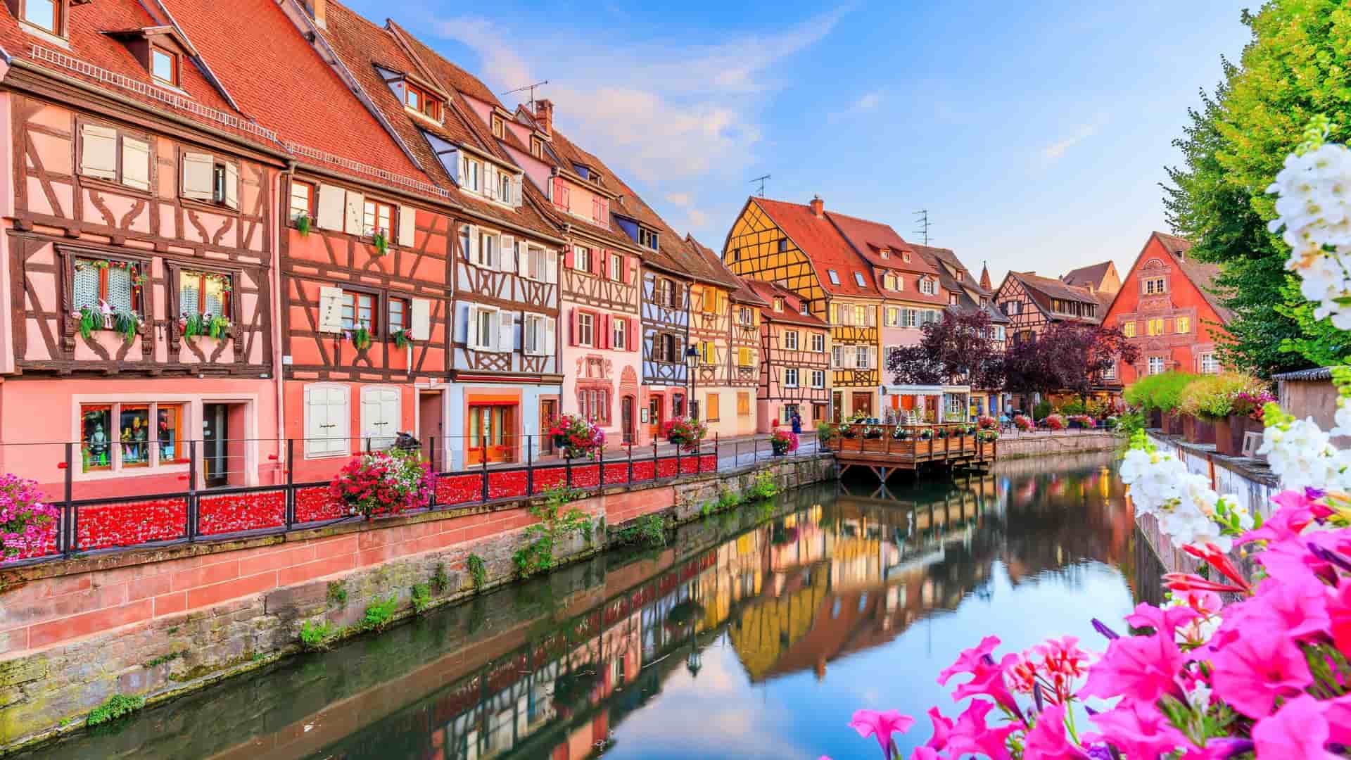 A picturesque canal scene in Colmar, France, showing a row of colorful, half-timbered houses with their reflections in the tranquil water, with vibrant flowers in the foreground.