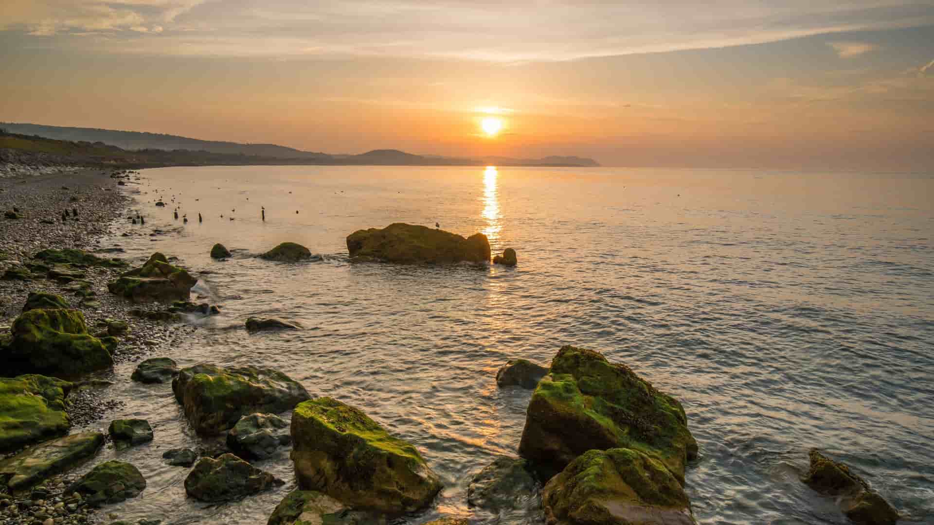 A serene sunset over the sea at Colwyn Bay, Wales, UK, illuminates mossy rocks on the shingle beach, with coastline hills in the distance.