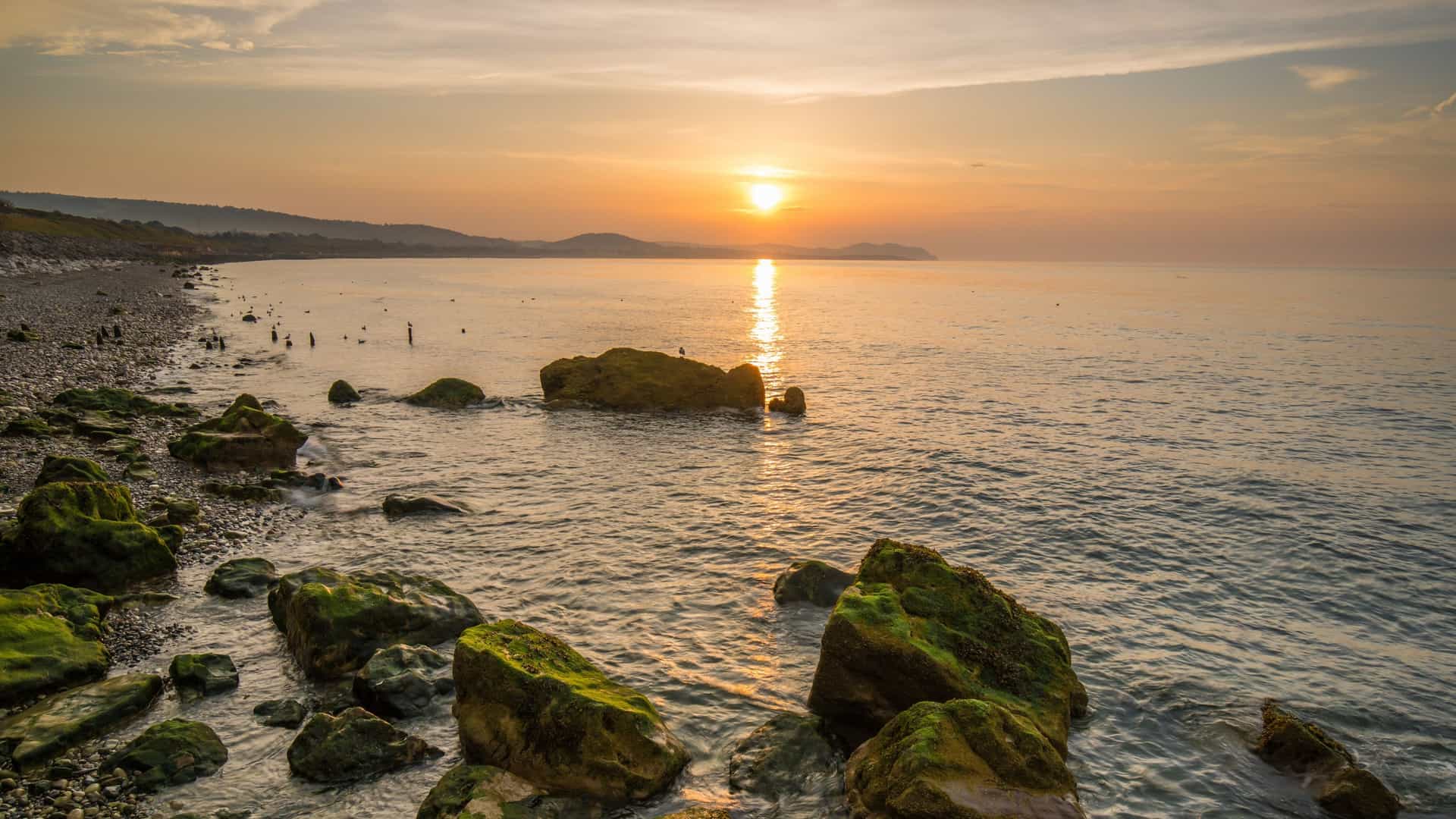 A serene sunset over the sea at Colwyn Bay, Wales, UK, illuminates mossy rocks on the shingle beach, with coastline hills in the distance.