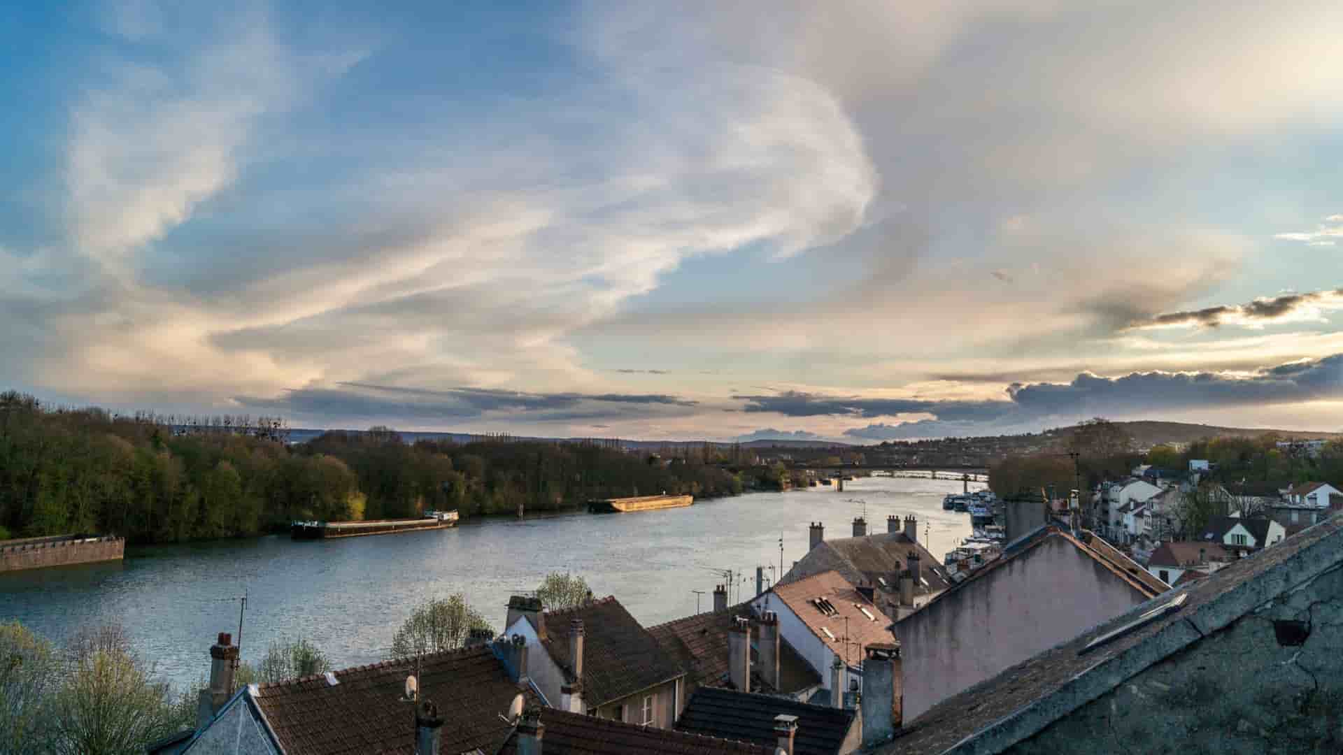 A picturesque view of the Seine River near Conflans-Sainte-Honorine, France, with rooftops of a charming village in the foreground and a barge sailing on the river.