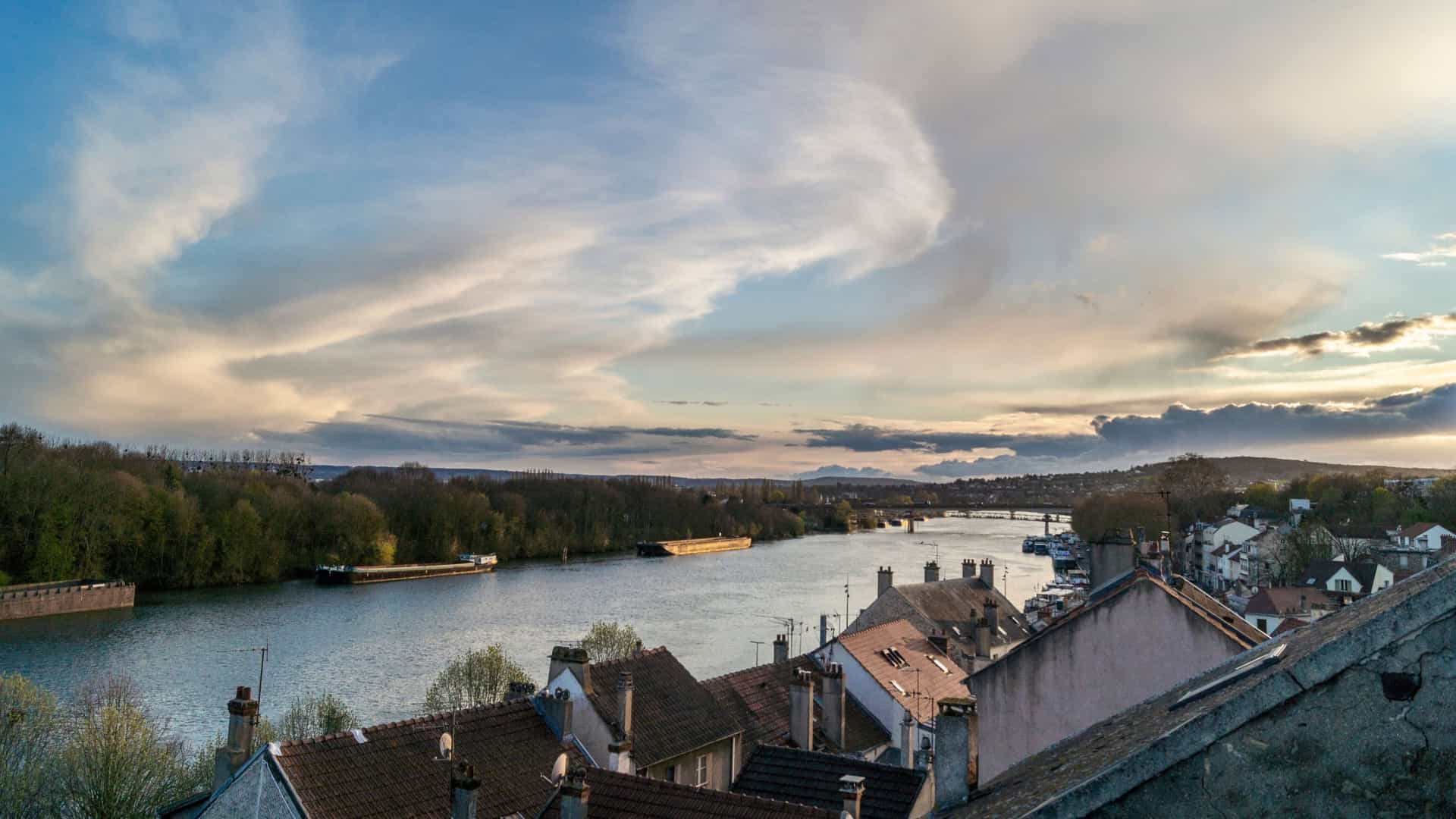 A picturesque view of the Seine River near Conflans-Sainte-Honorine, France, with rooftops of a charming village in the foreground and a barge sailing on the river.
