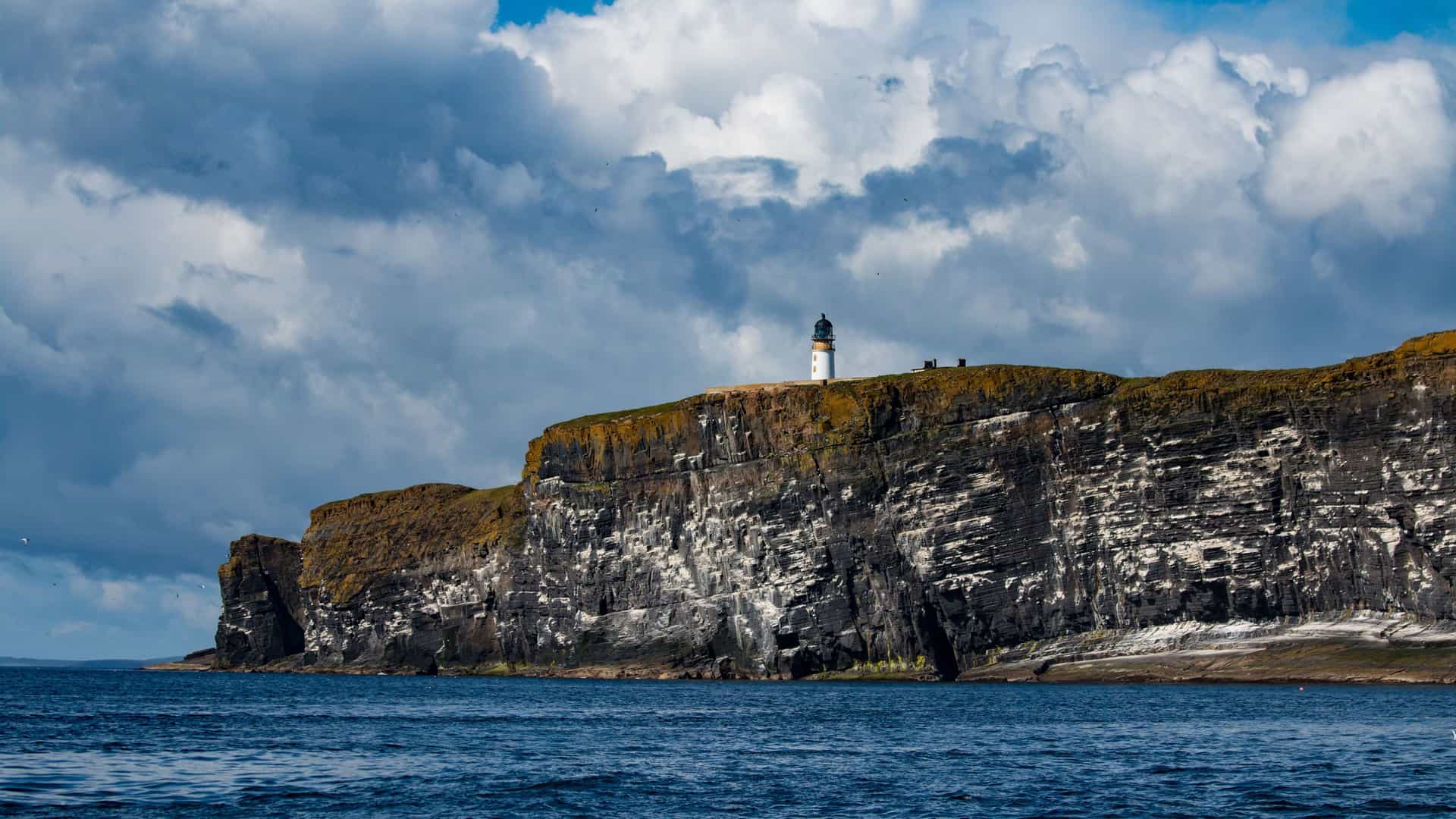A white lighthouse perched atop the dramatic, sheer cliffs of Copinsay, one of the remote Orkney Islands, with a vast blue sea and cloudy sky.