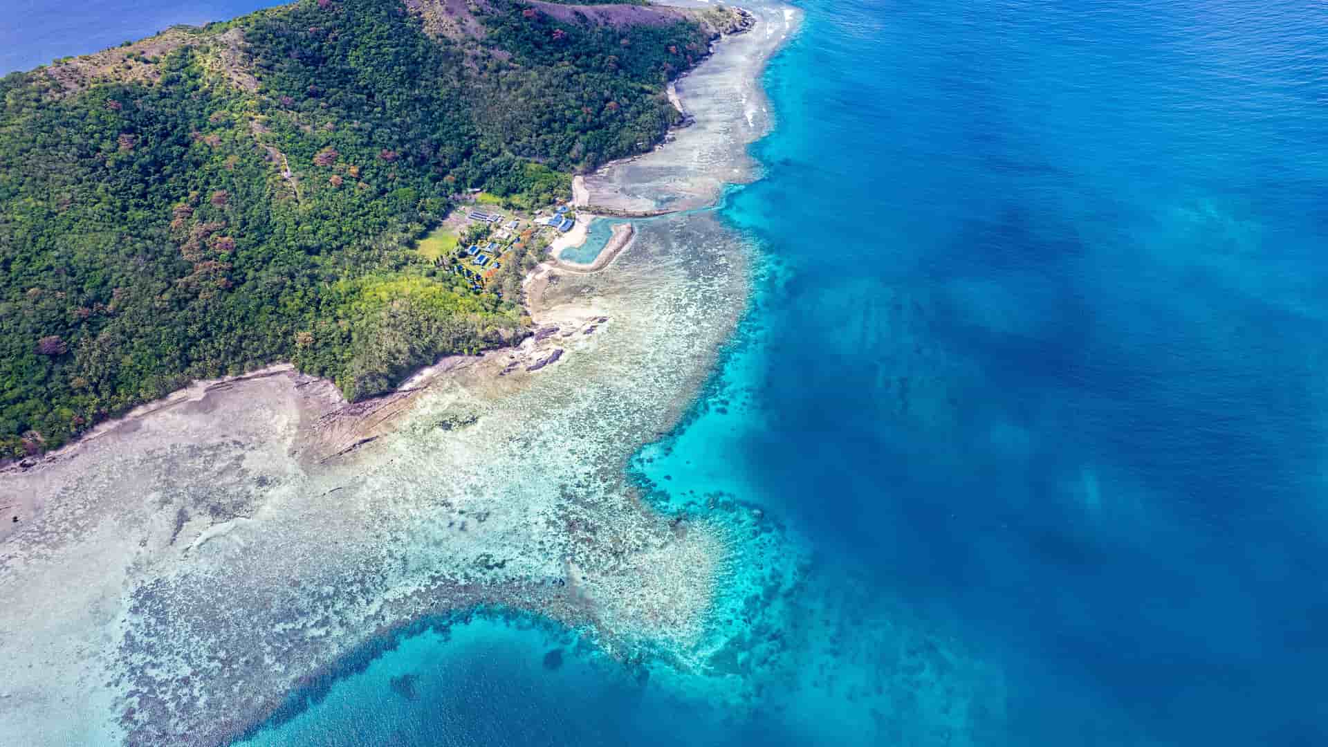"An aerial view of the stunning Coral Coast in Fiji, showing a lush green island meeting the vibrant blue water of the Pacific Ocean, with a shallow coral reef visible beneath the surface. "
