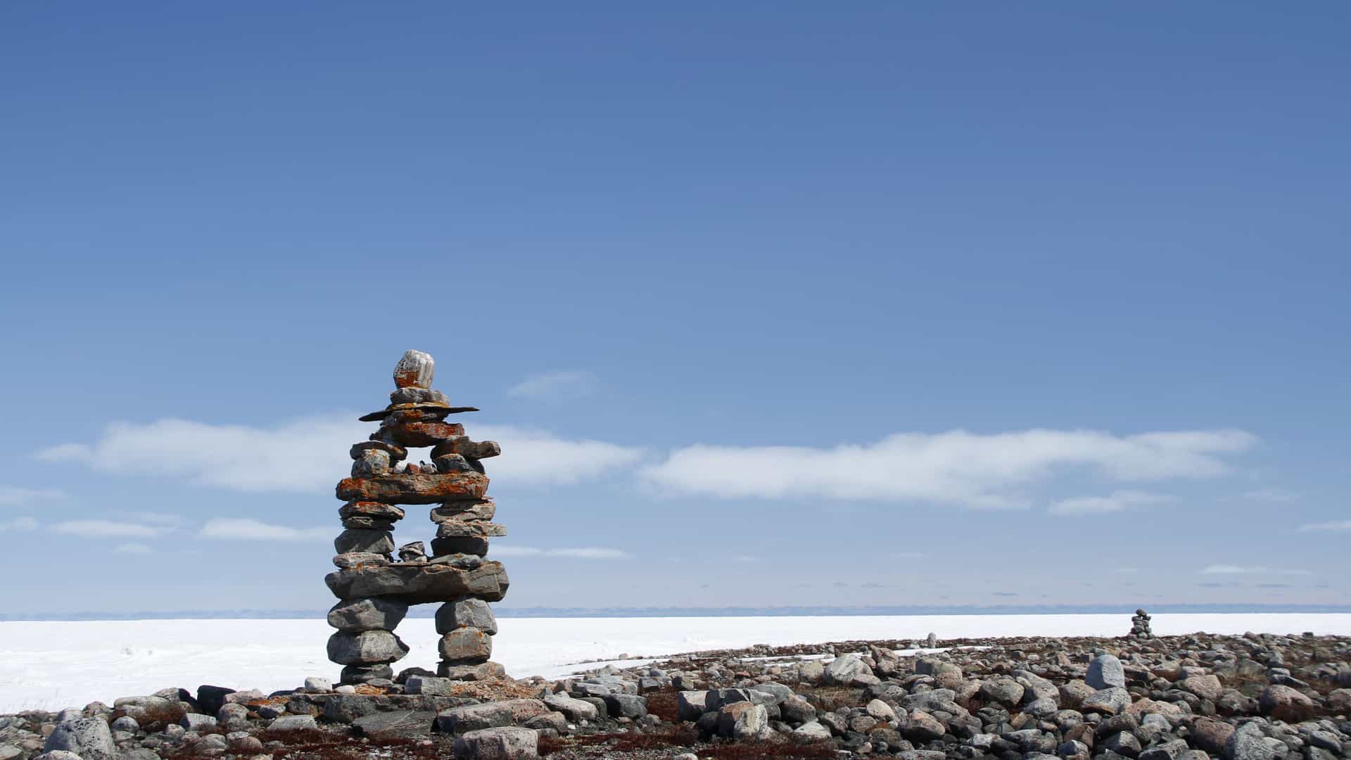 A tall, stone inuksuk stands on a rocky shore near Coral Harbour, Nunavut, with a vast, frozen expanse and a light blue sky with wispy clouds.