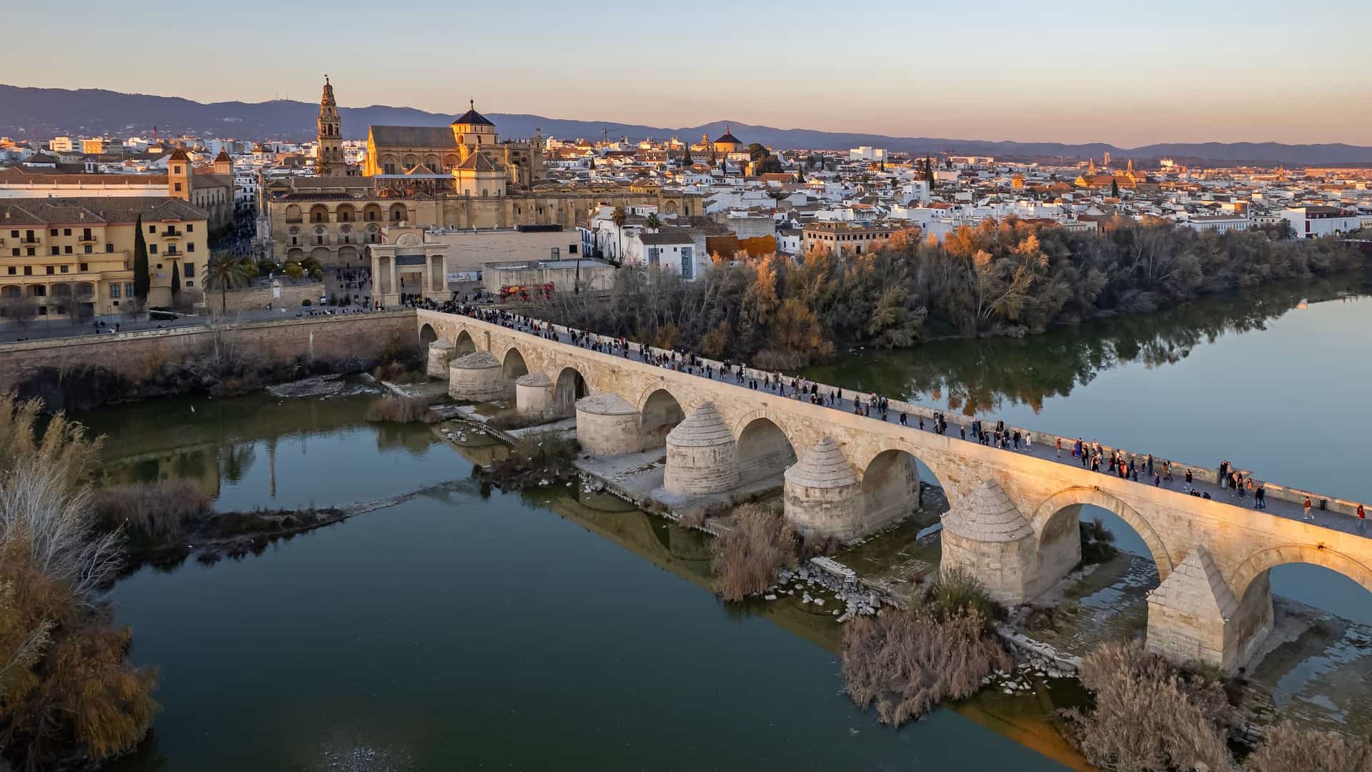 "An aerial view of the historic Roman Bridge of Córdoba, Spain, with tourists walking across its many arches over the Guadalquivir River, leading to the ancient Mezquita-Cathedral and the city skyline during sunset. "