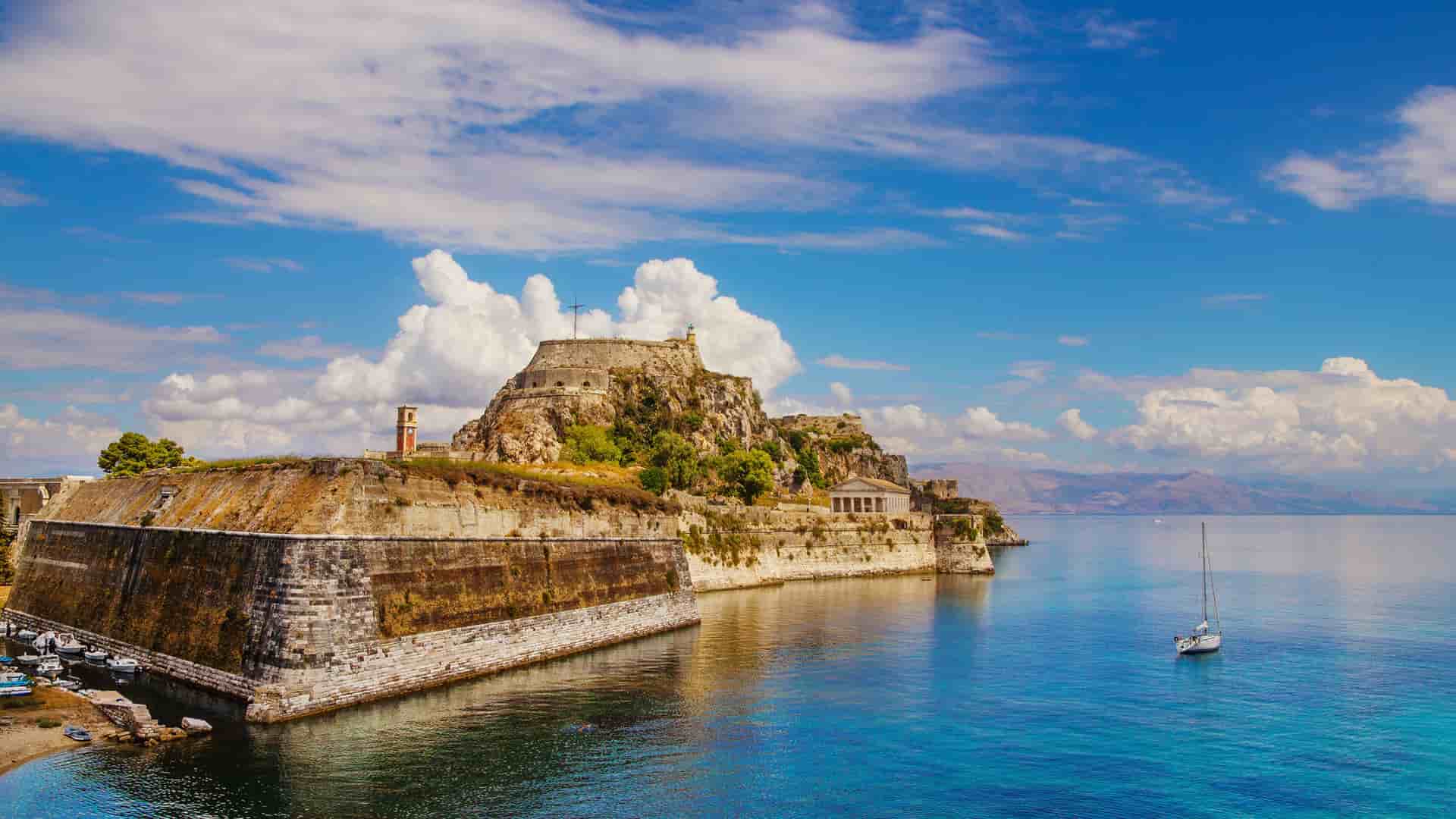 A picturesque view of the historic Old Fortress of Corfu, Greece, with its stone walls rising from the turquoise Ionian Sea and a sailboat in the foreground.