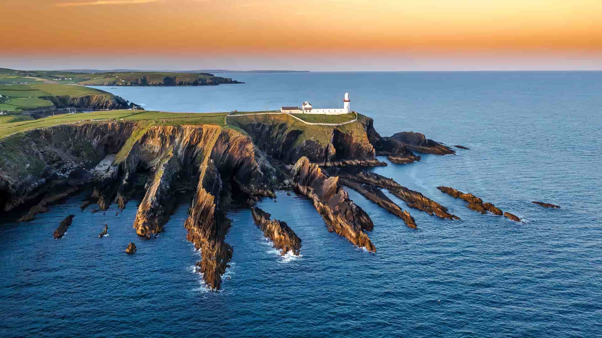 A scenic view of the white Galley Head Lighthouse perched on the dramatic, jagged cliffs of the West Cork coastline in Ireland at sunset.