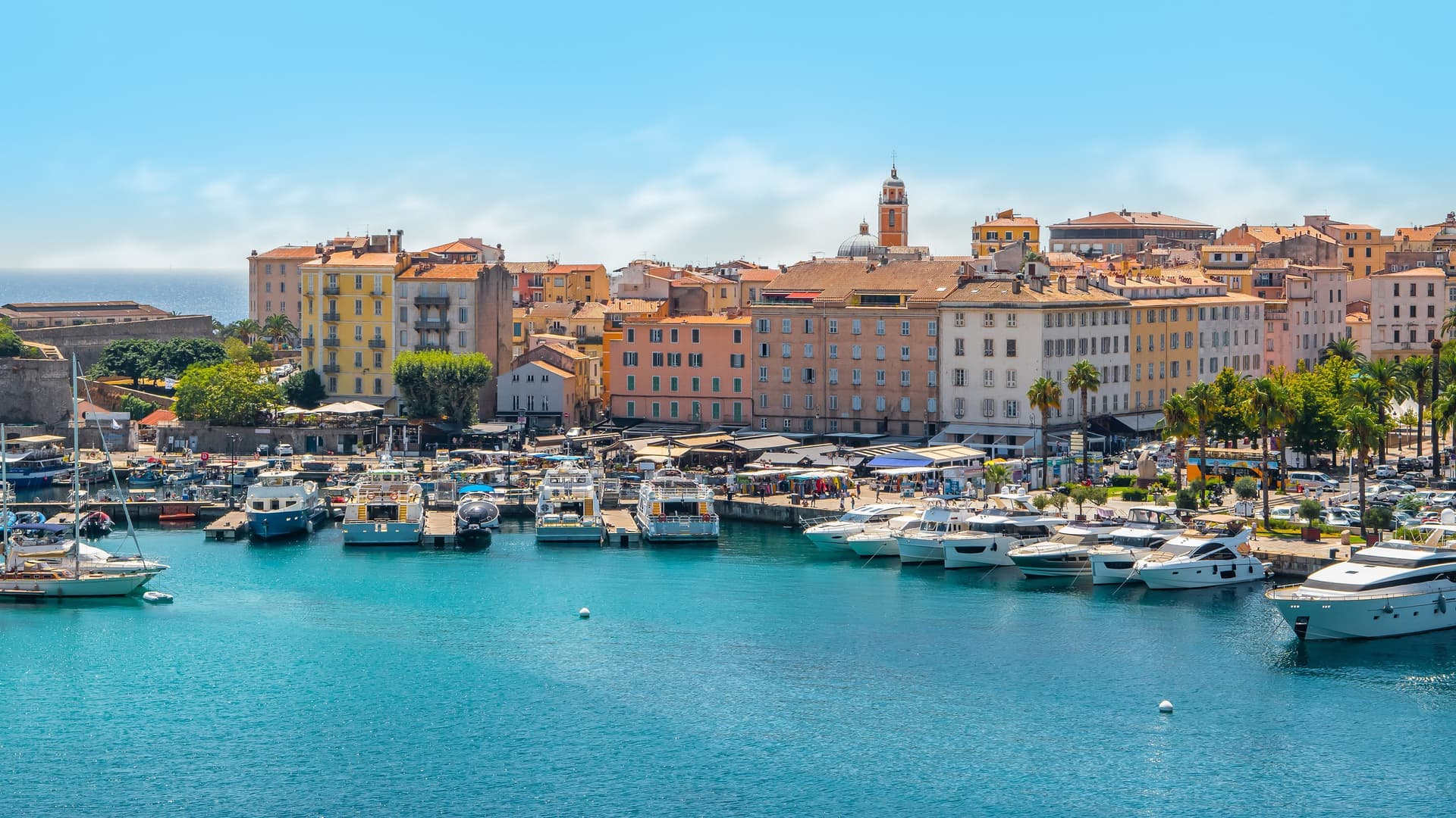 A beautiful view of the bustling harbor in Ajaccio, Corsica, with colorful historic buildings lining the port and numerous yachts docked in the marina.