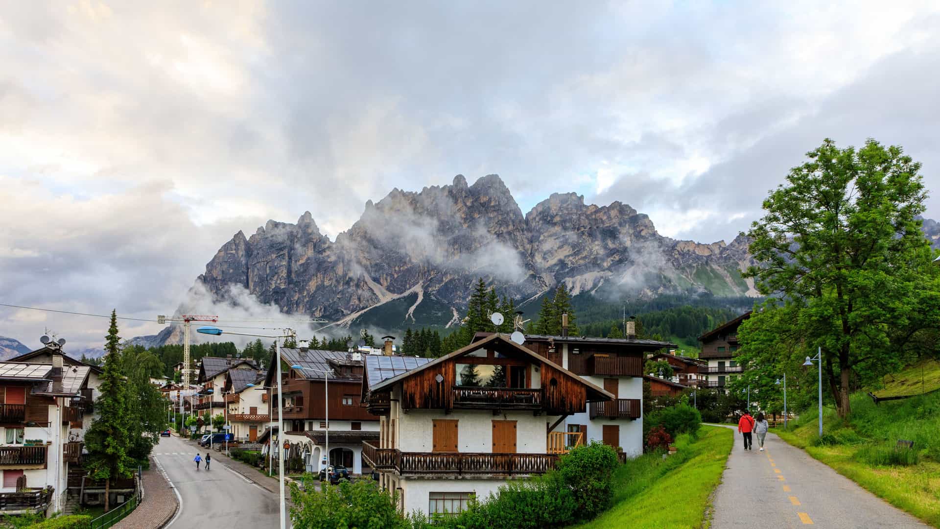 "A picturesque village scene in Cortina d'Ampezzo, Italy, with charming alpine buildings and a couple of people walking on a path, set against the dramatic, rocky peaks of the Dolomite mountains under a cloudy sky. "