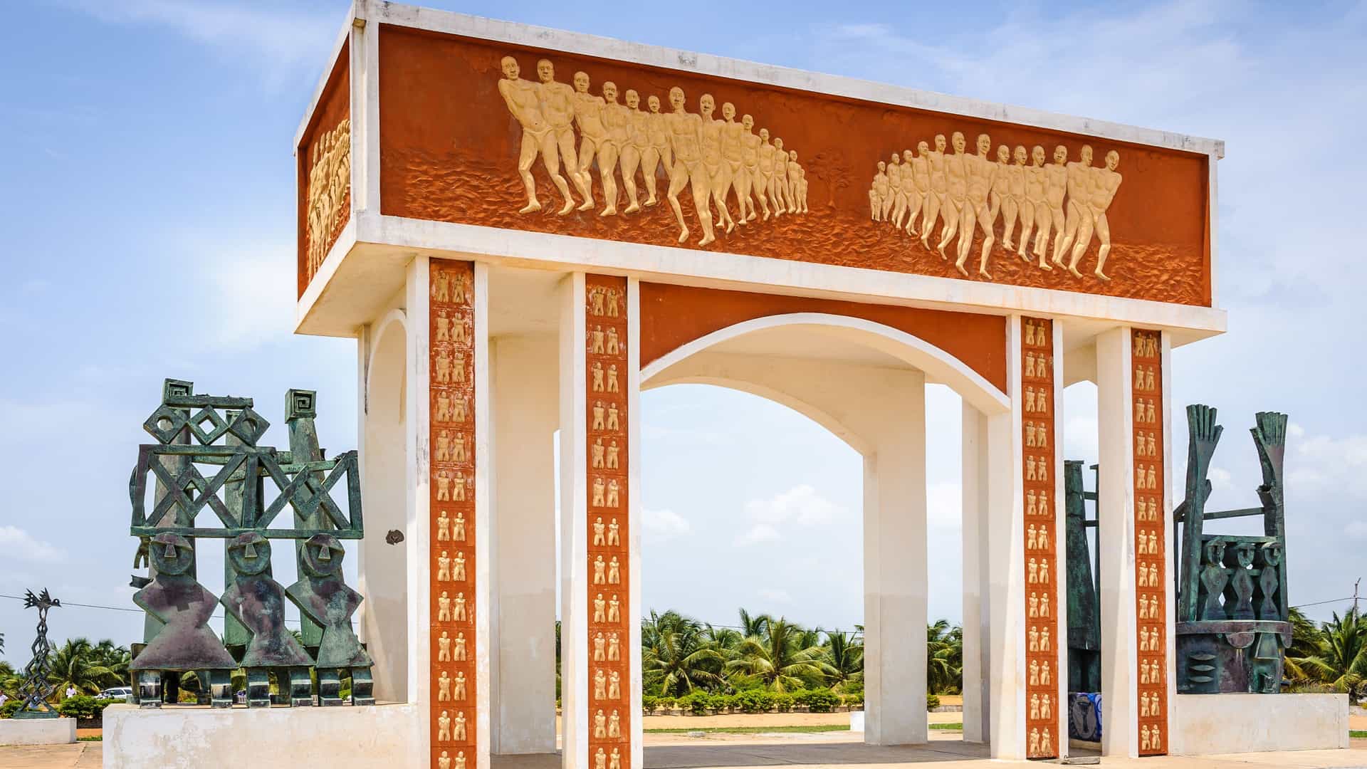 A unique public monument in Cotonou, Benin, featuring a large archway with vibrant relief carvings of people and abstract sculptures on either side.
