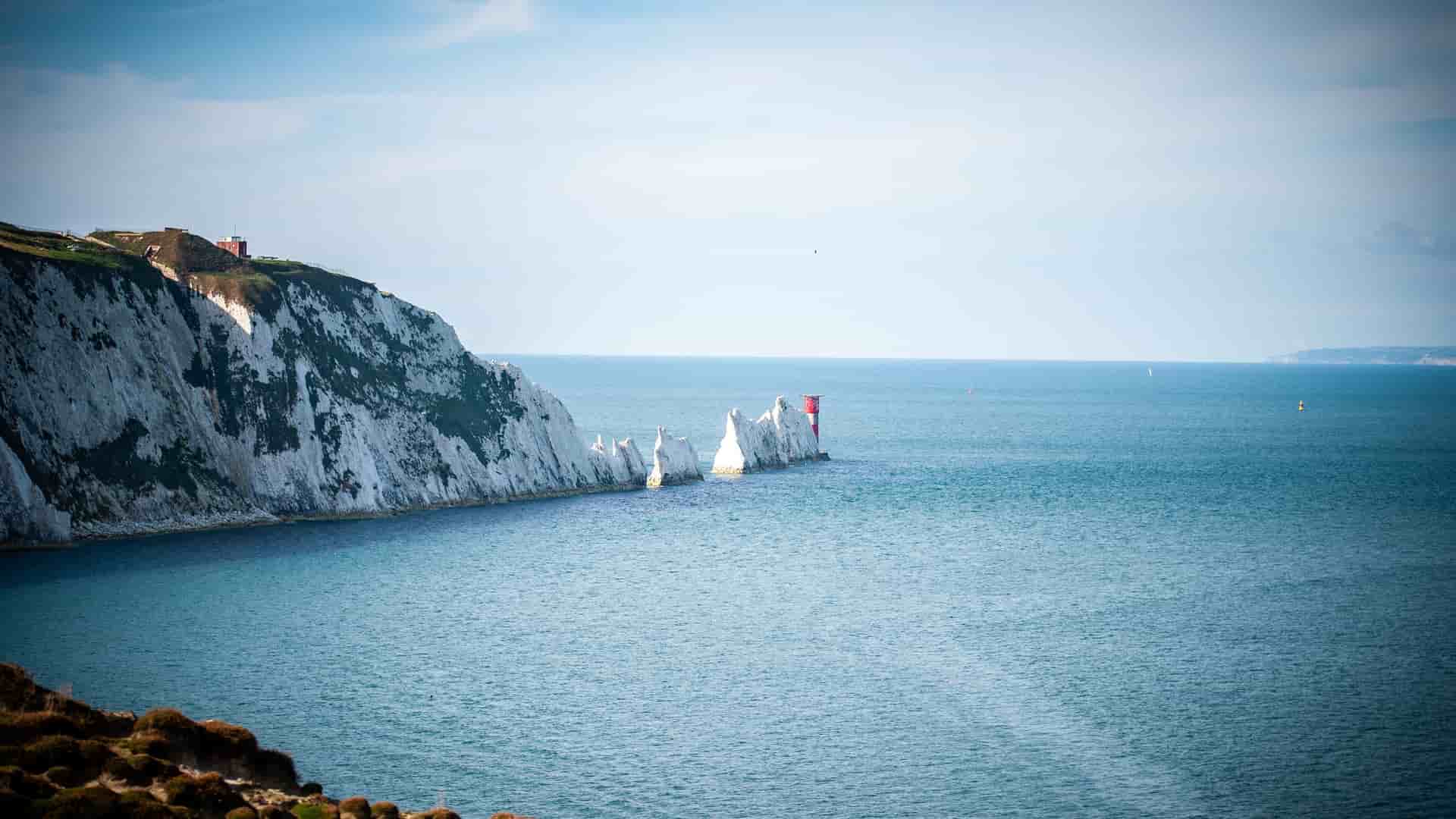 A scenic view of The Needles, iconic chalk stacks with a red and white striped lighthouse, extending from the white cliffs of the Isle of Wight into the blue sea.
