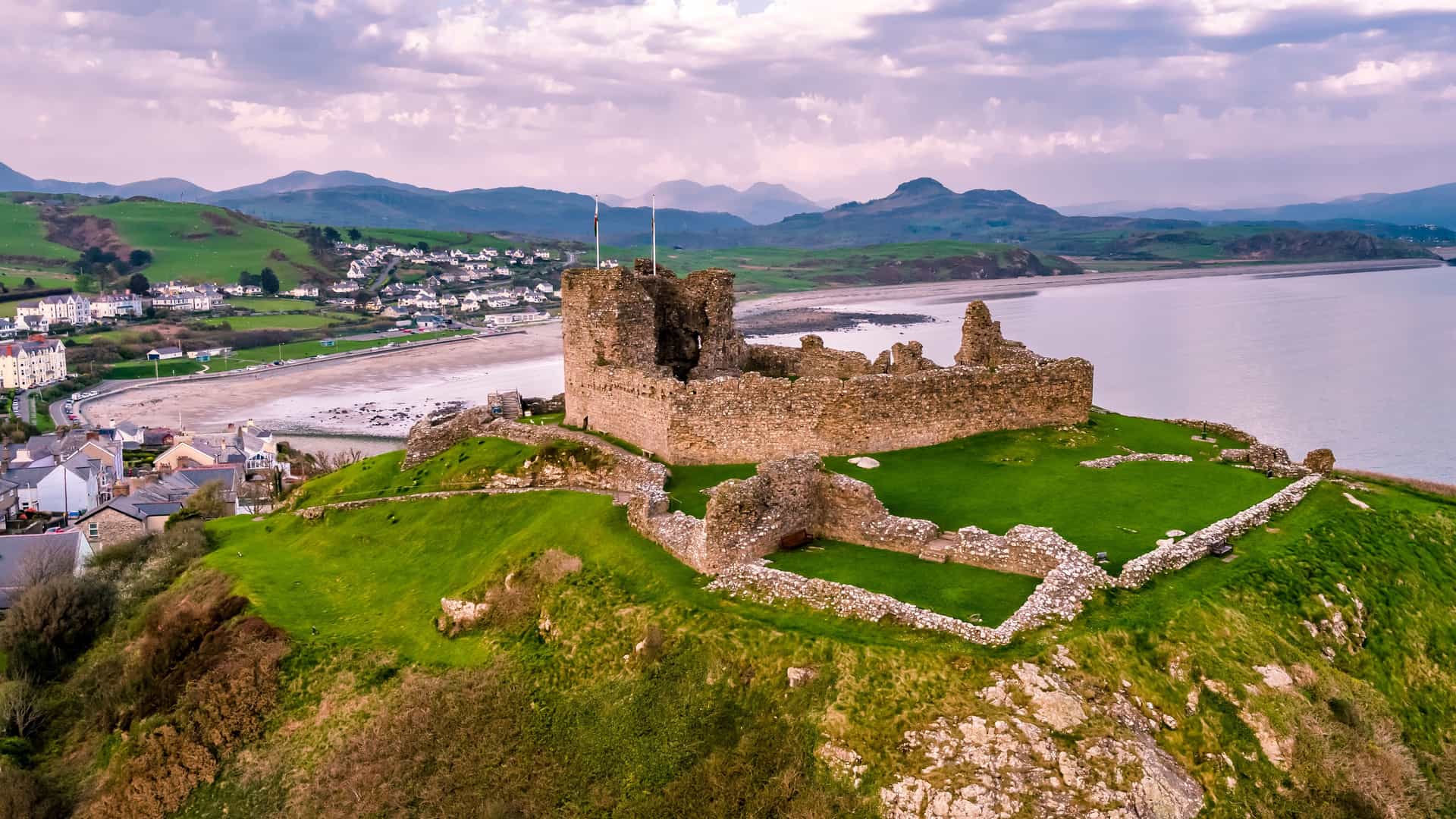 "An aerial view of the historic Criccieth Castle ruins, with its stone walls sitting on a grassy headland, overlooking a sandy beach and the Irish Sea in Wales, with a small town and green hills in the background. "