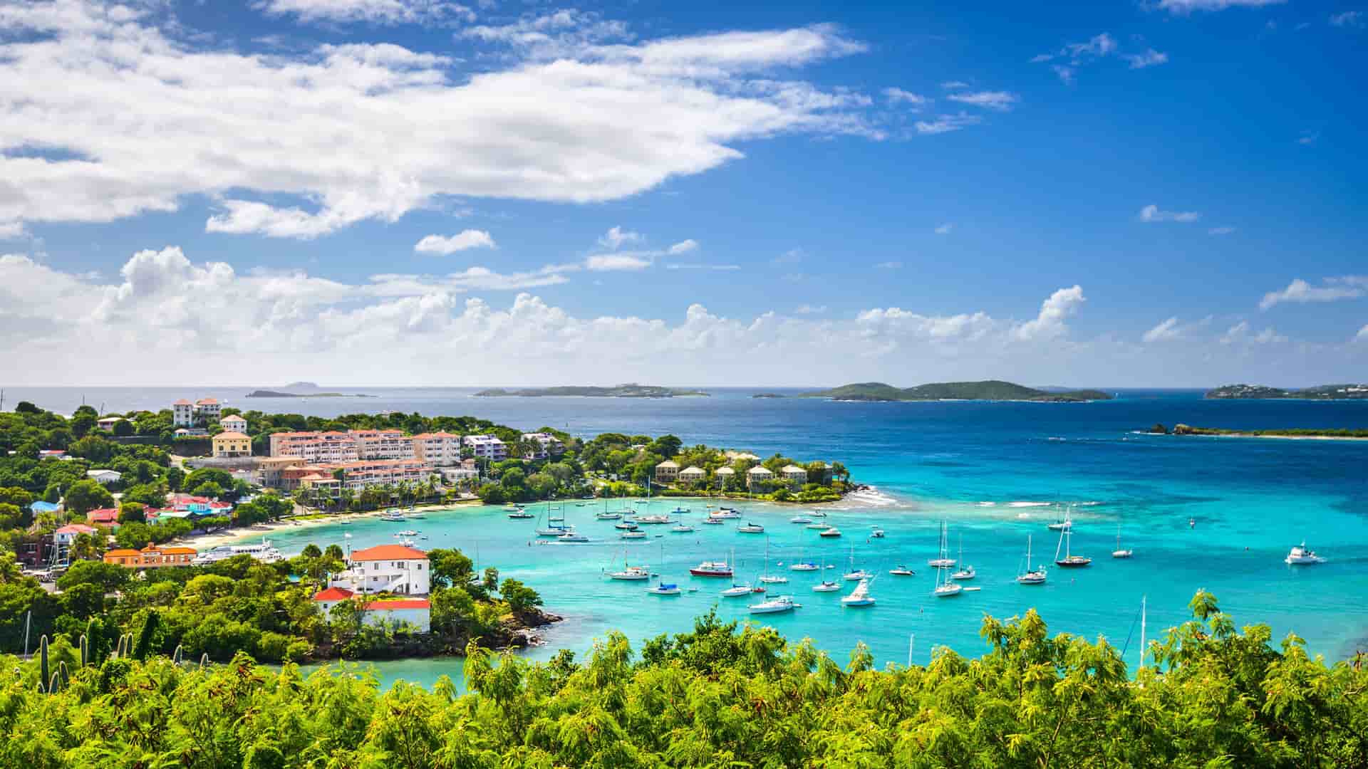 A breathtaking panoramic view of Cruz Bay, St. John, showcasing a vibrant coastal town with white-sand beaches, clear turquoise waters, and numerous boats in the harbor.