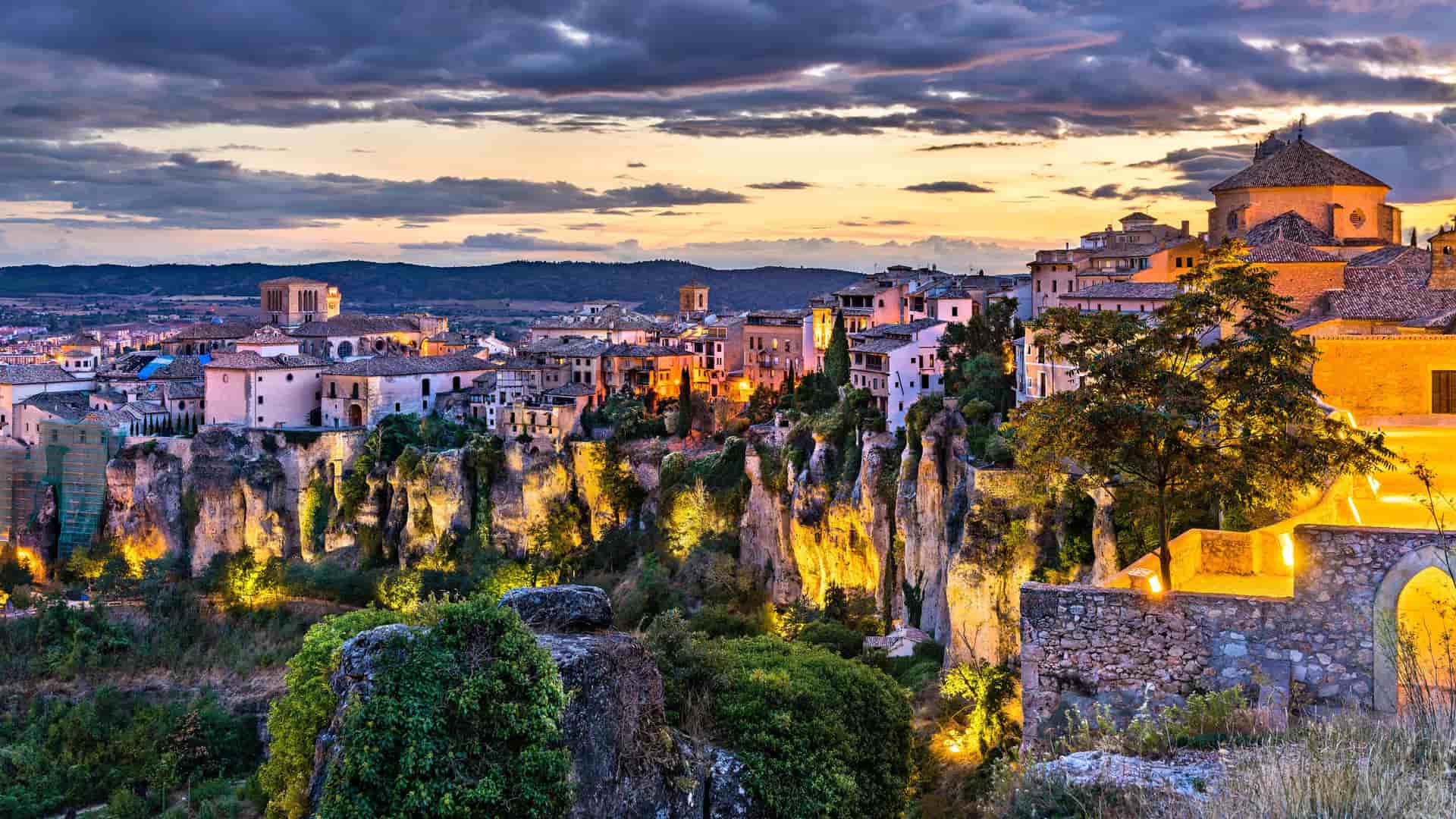 "A breathtaking image of the historic city of Cuenca, Spain, at dusk, showing the famous ""Hanging Houses"" built precariously on the cliffs, illuminated against the dramatic sunset sky. "