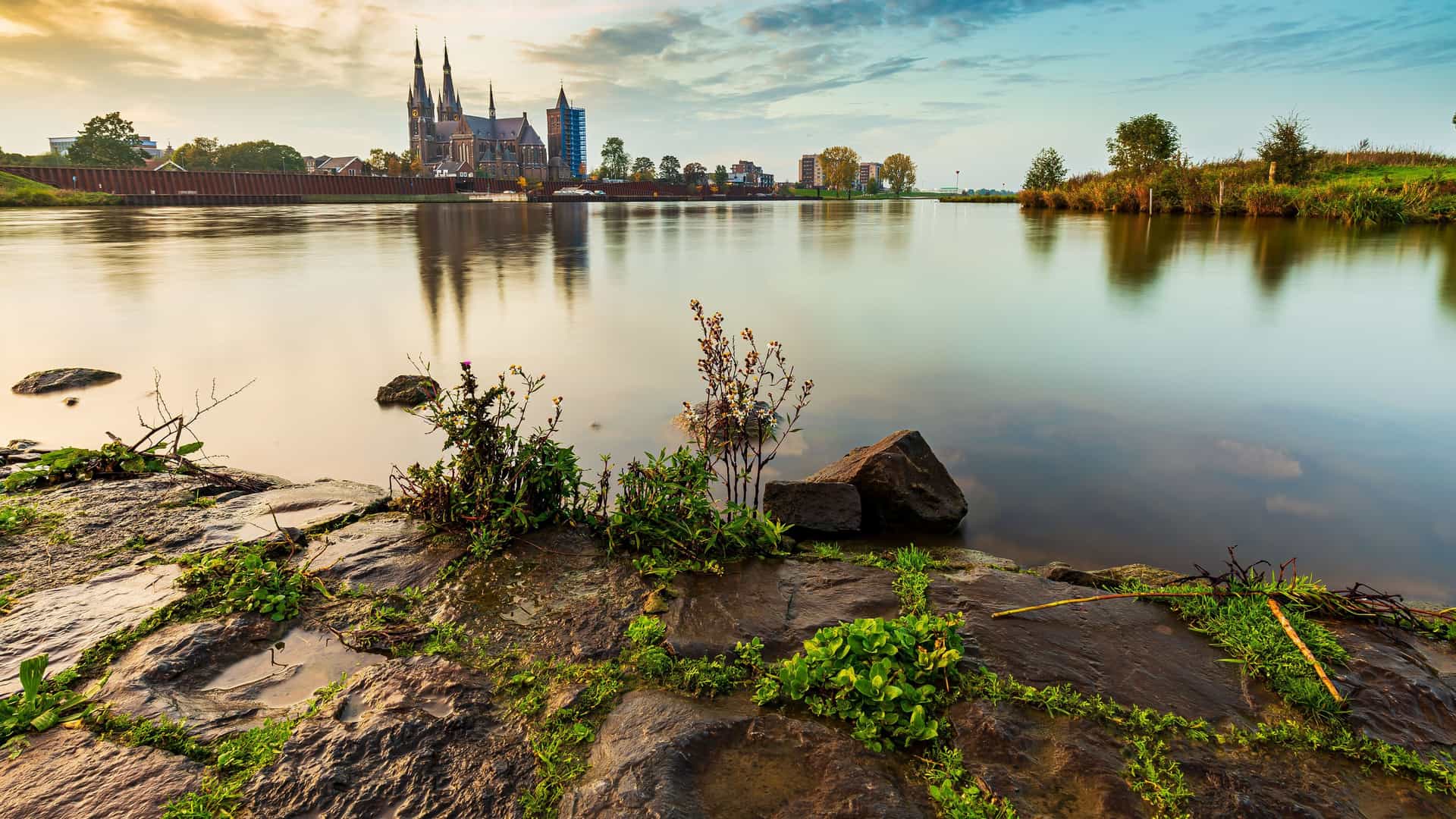 A scenic view of the St. Martin's Church in Cuijk, Netherlands, reflecting on the calm Meuse River during a beautiful sunset, as seen from the rocky shoreline.